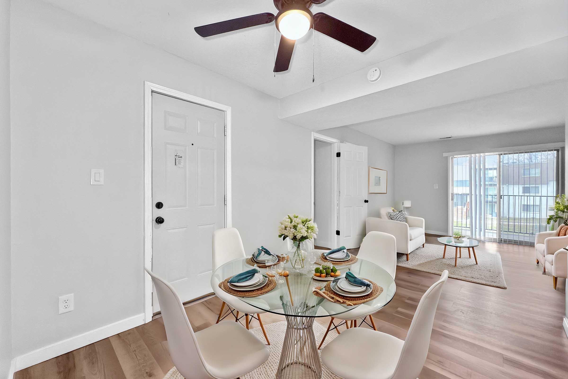 Bright and airy living space featuring a dining area with a glass table set for four. White chairs surround the table, and a small vase of flowers adds a decorative touch. The room has light-colored walls, a ceiling fan, and large windows letting in natural light. A cozy seating area is visible in the background.