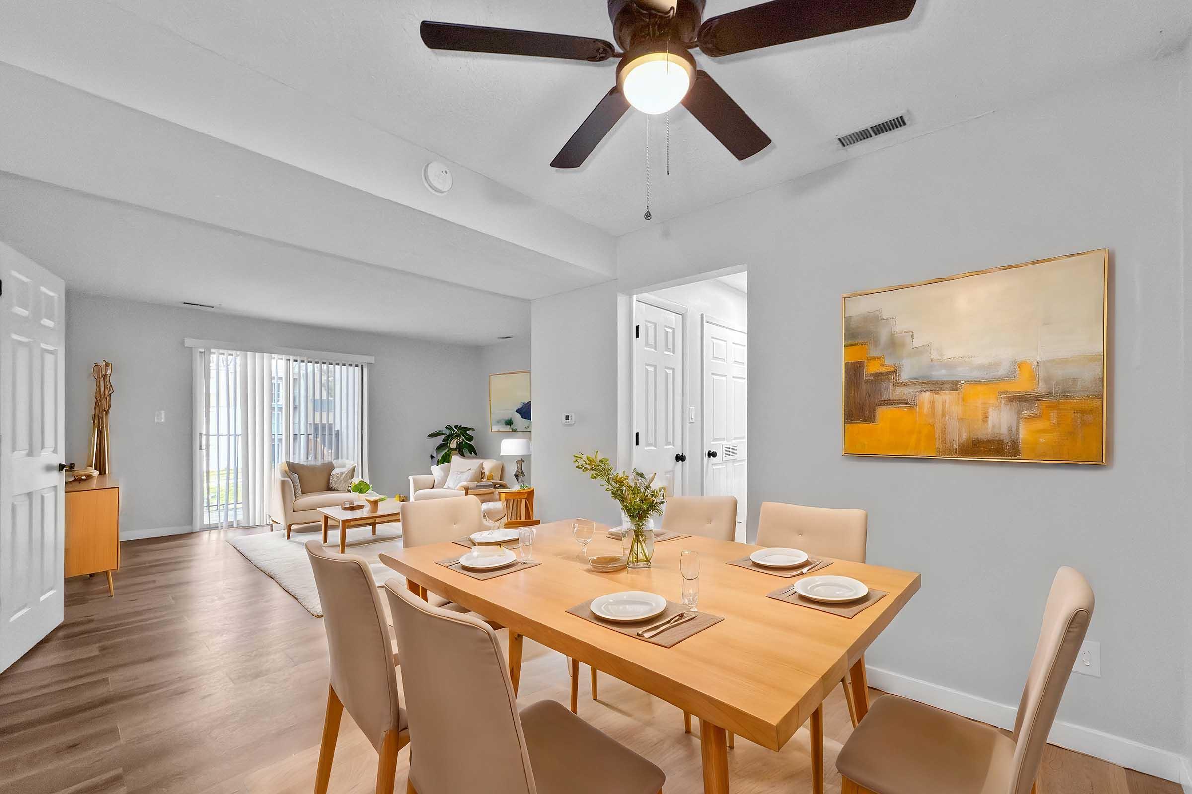 A modern dining area featuring a wooden table set for four with plates and glasses. Light-colored chairs surround the table. In the background, a cozy living room is visible with soft seating and large windows. A wall artwork in warm tones hangs near the dining area, complemented by a ceiling fan.