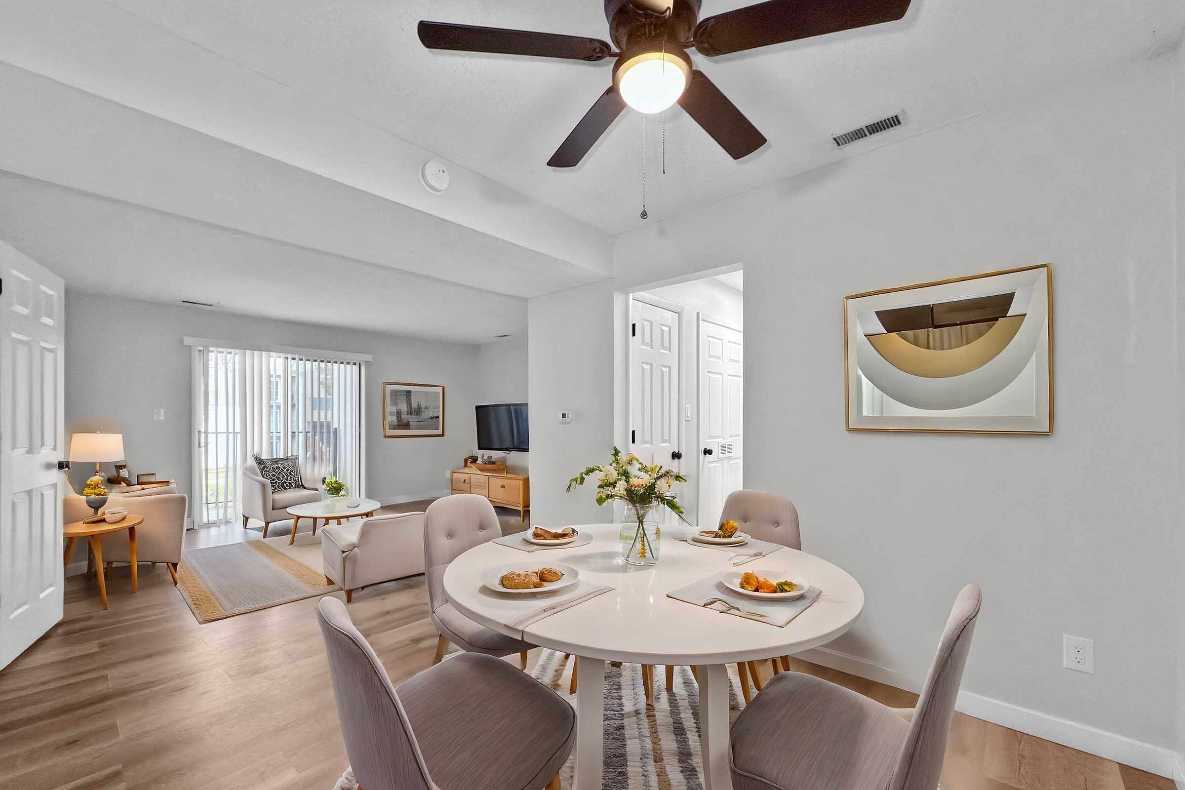 A modern dining area with a round table set for four, featuring plates of food and a vase of flowers. The background includes a living space with a sofa and a TV, and a doorway leading to another room. Natural light streams in through sliding glass doors.