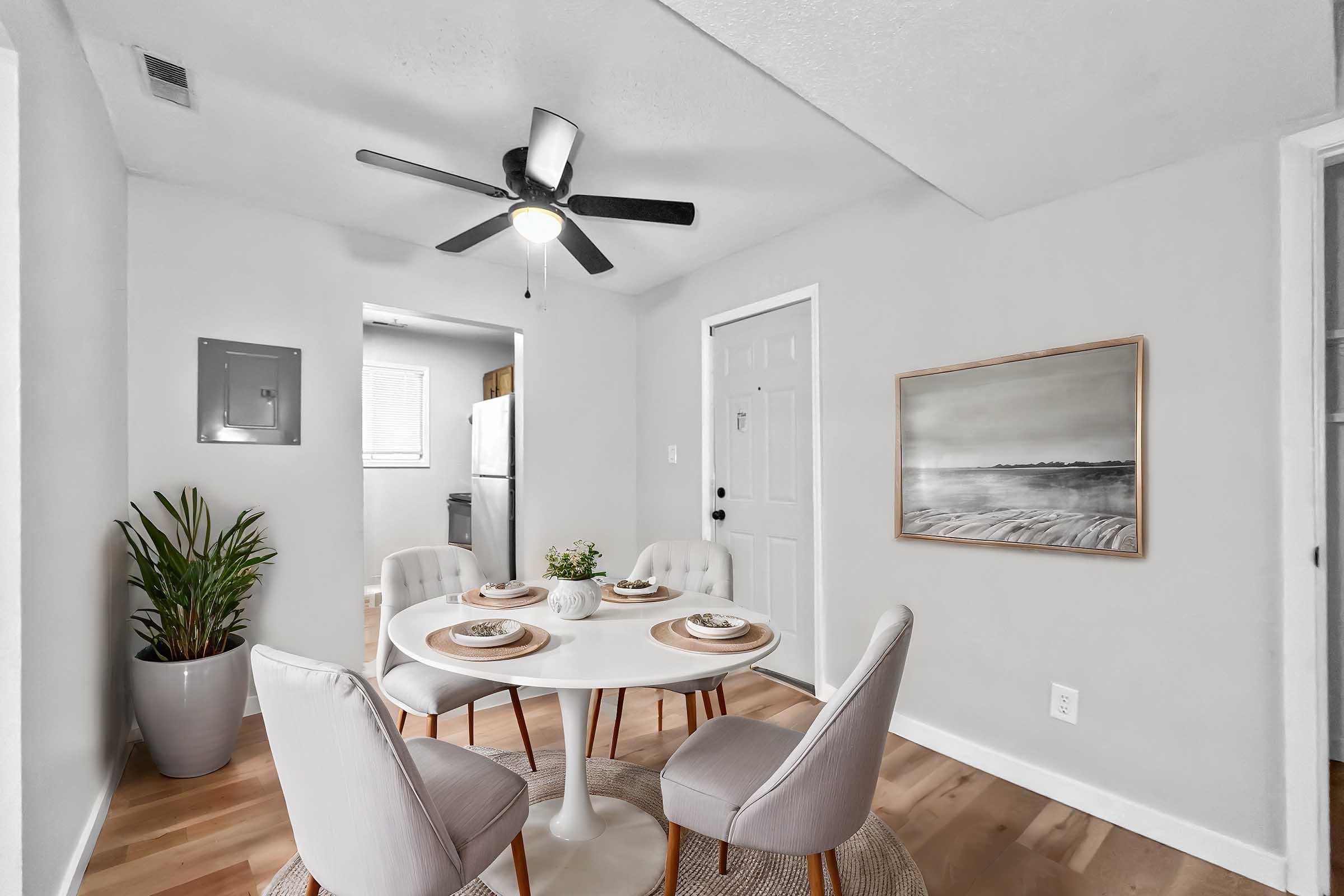 A modern dining area featuring a round white table set for four with elegant tableware. The room has light gray walls, a ceiling fan, and a large plant in a pot. Visible in the background is a door leading to another room. A piece of art hangs on the wall, depicting a serene landscape.