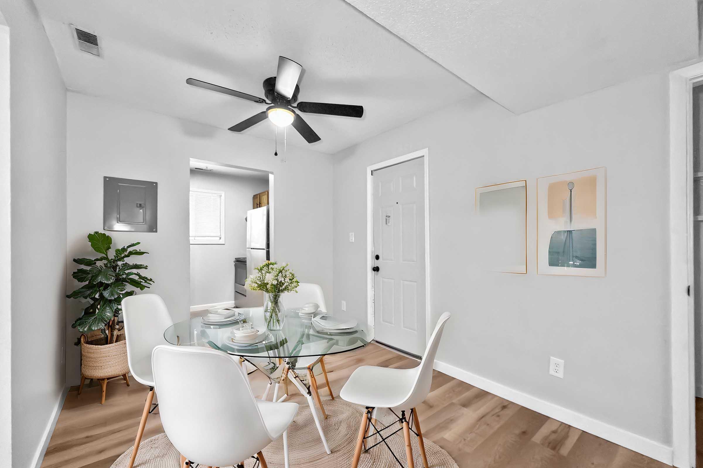A modern dining area featuring a glass table set for four with white chairs. A potted plant adds greenery to the space. The walls are painted light gray, and there is a ceiling fan above. A door leads to another room, and there are two framed artworks hung on the wall.