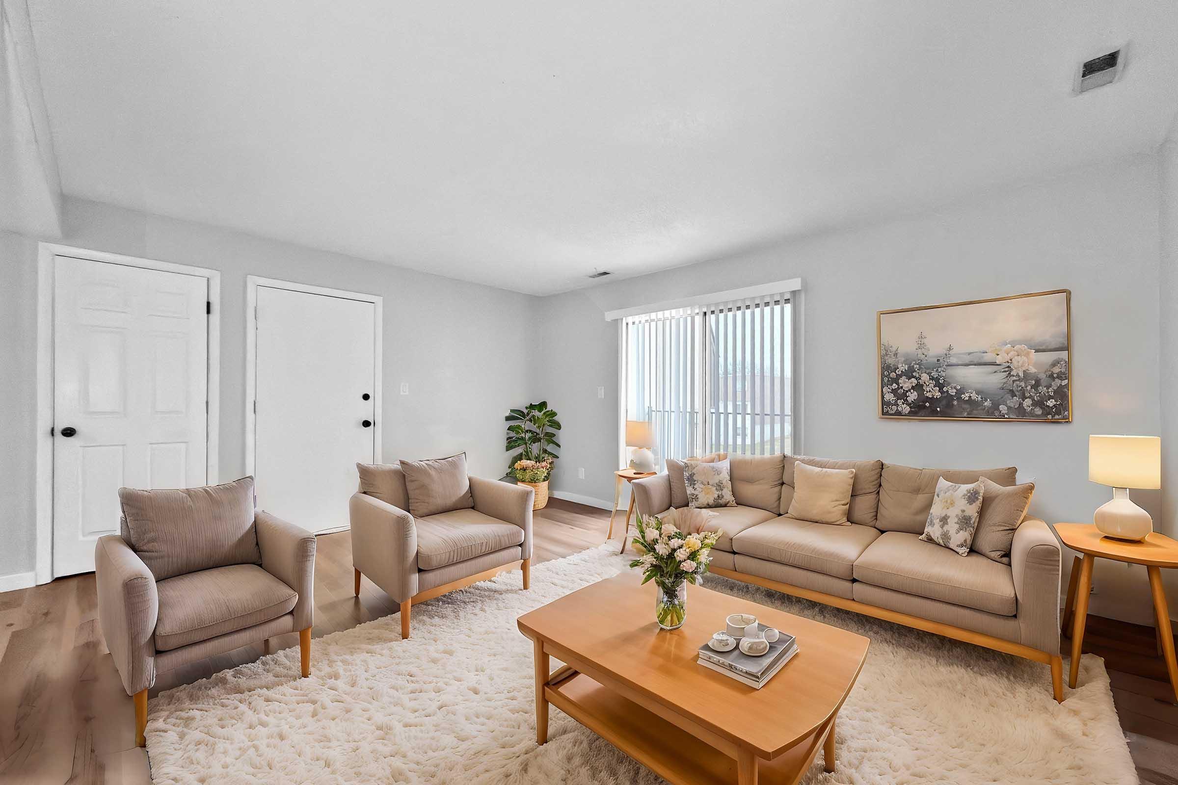 A cozy living room featuring a light beige sofa with decorative pillows, two matching armchairs, and a wooden coffee table. A floral arrangement is placed on the table. The room has neutral walls, large windows with sheer curtains, and a soft area rug, creating a welcoming and bright atmosphere.