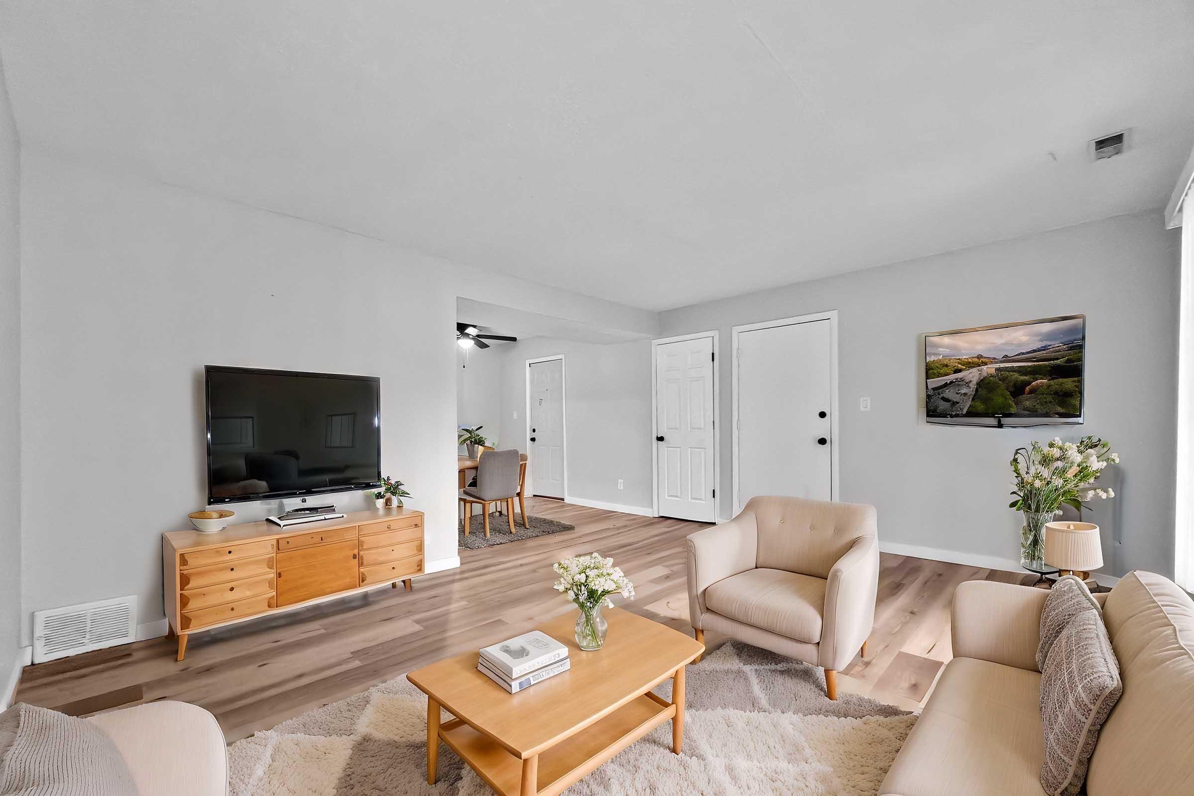 A modern living room featuring a light color scheme. It includes two beige armchairs, a wooden coffee table with a vase of flowers, and a TV on a wooden stand. A wall-mounted television displays a nature scene, and there are double doors leading to another room. Bright, inviting atmosphere with natural light.
