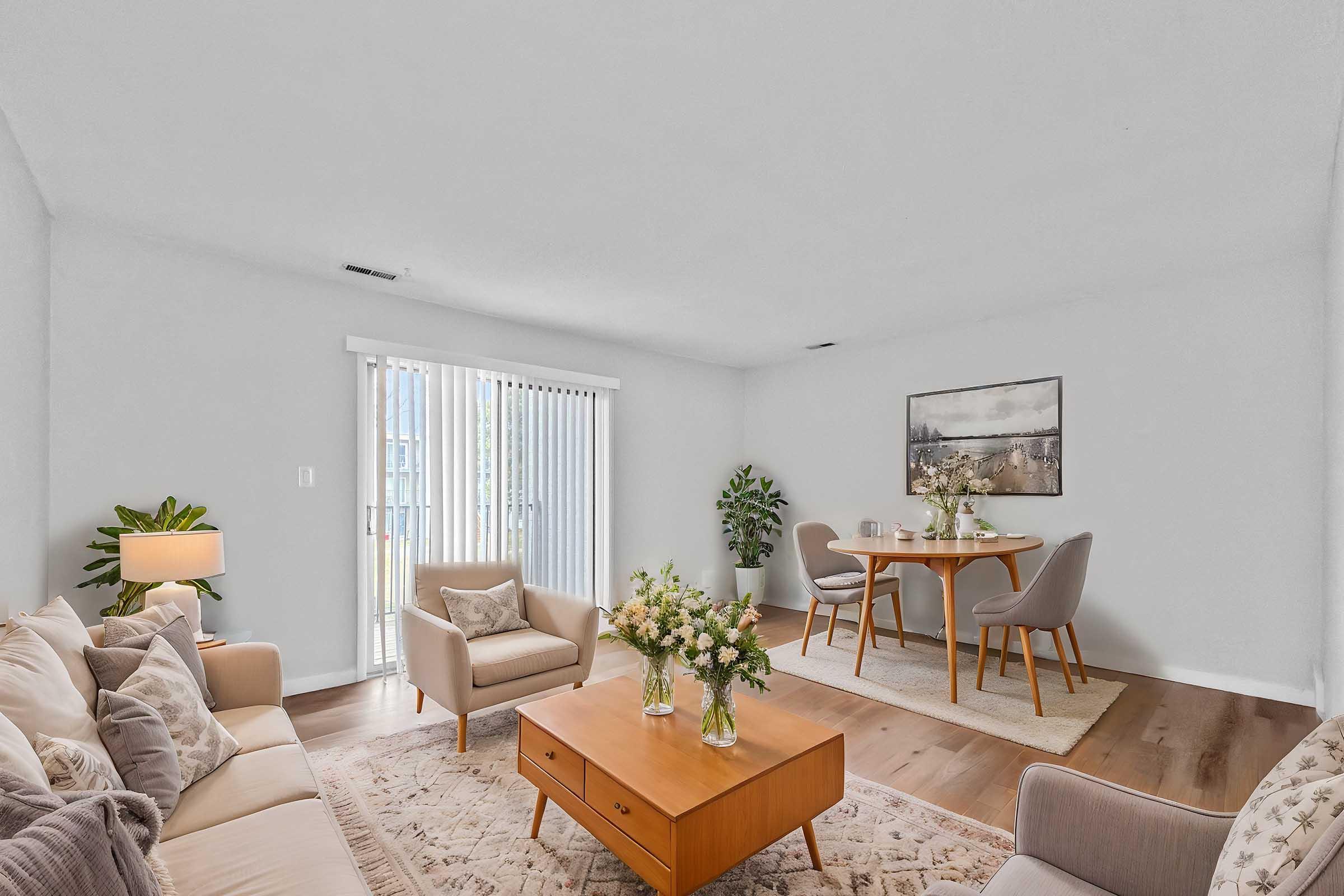 A modern, light-filled living room featuring a beige sofa and armchairs, a wooden coffee table with a vase of flowers, and a cozy dining area with a round table and chairs. Large sliding glass doors lead to a patio, and there are potted plants and neutral decor throughout the space.