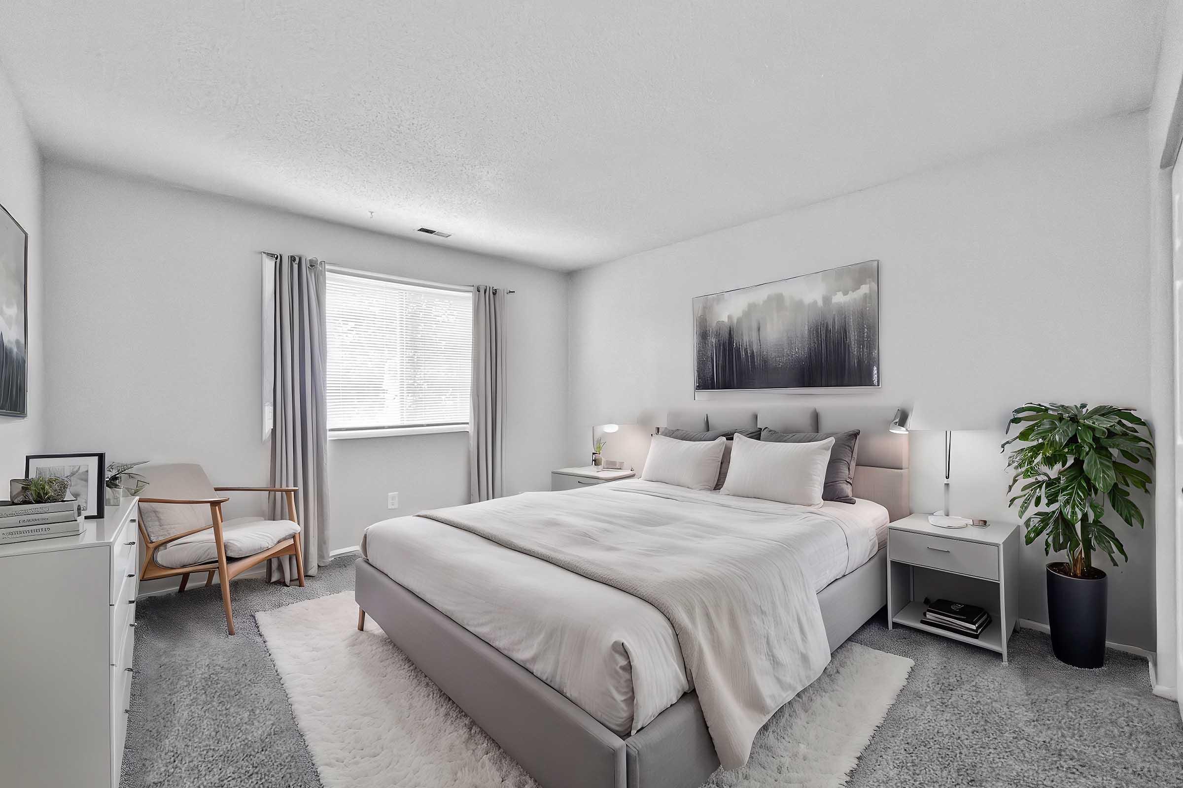 A modern bedroom featuring a queen-sized bed with neutral bedding, a stylish chair, and a green potted plant. Soft gray walls and carpet create a cozy atmosphere, complemented by curtains and a piece of abstract wall art. The room includes a nightstand, lamp, and window allowing natural light.