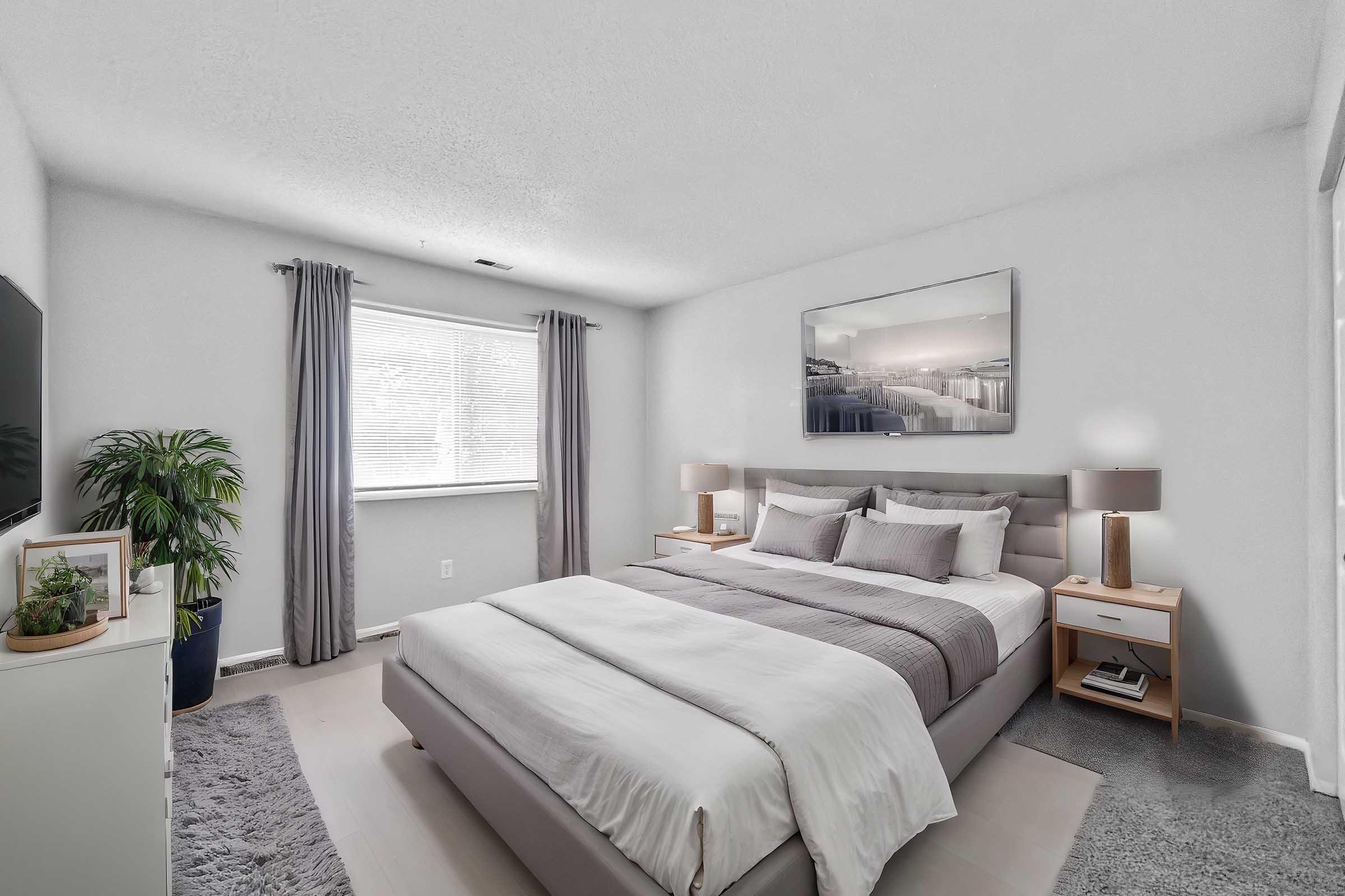 A modern bedroom featuring a stylish gray bed with white and gray bedding, two nightstands with lamps, a large window with sheer curtains, and a decorative plant in the corner. The walls are painted light gray, and a framed abstract artwork hangs above the bed, creating a serene atmosphere.