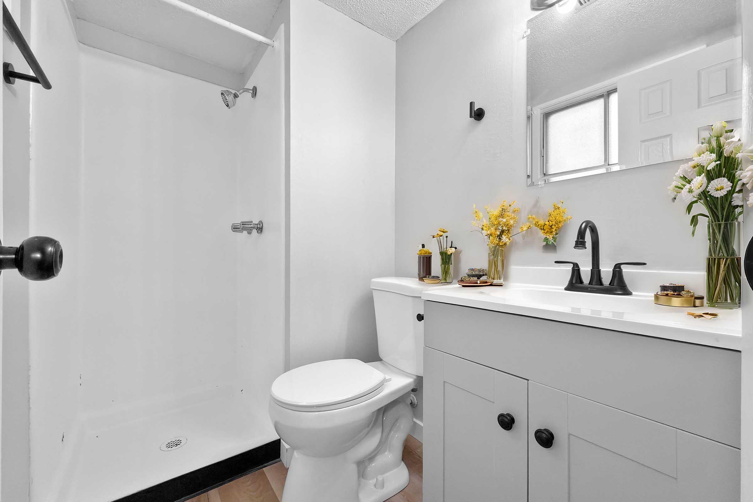 A clean, modern bathroom featuring a white shower stall, toilet, and a stylish vanity with a sink. The vanity has dark cabinet handles, decorative items on top, and a vase with yellow flowers. Bright lighting and neutral-colored walls enhance the spacious feel.