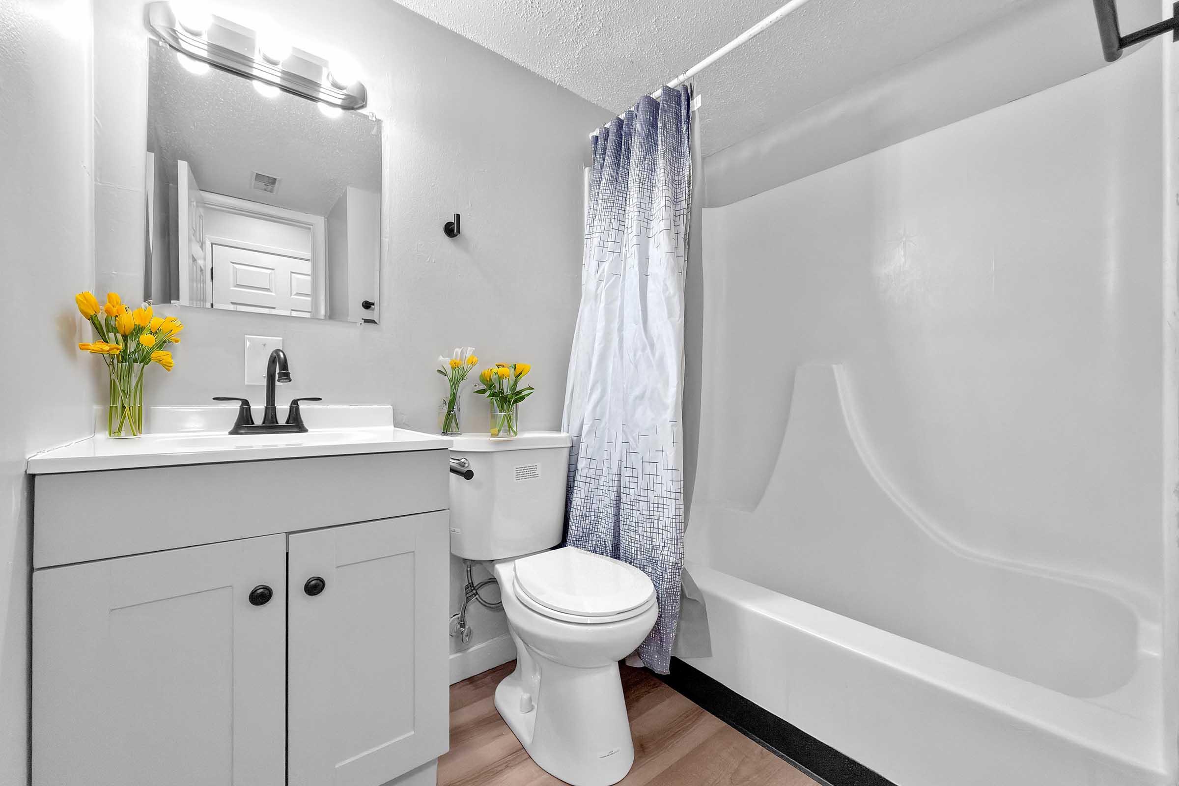 A bright and modern bathroom featuring a white bathtub with a shower, a toilet, a vanity with a sink, and a large mirror. Fresh yellow tulips are arranged on the vanity, adding a pop of color. The walls are light grey, and a patterned shower curtain complements the décor.