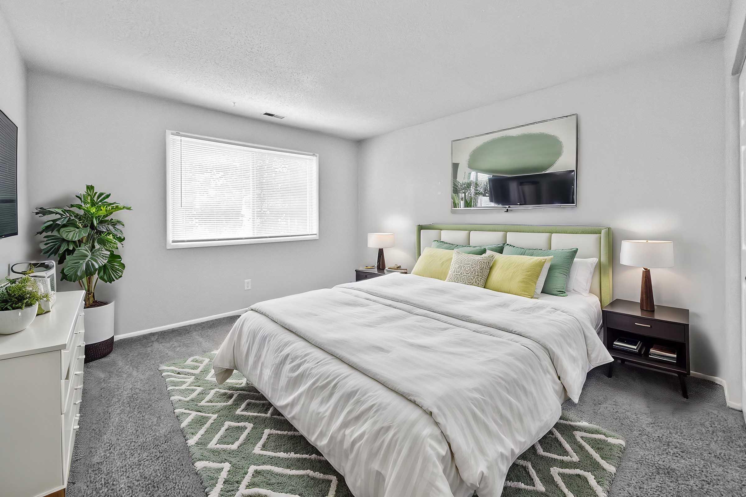 A well-decorated bedroom featuring a large bed with white linens and green accent pillows. There are two bedside lamps on matching nightstands, a large artwork above the bed, a green area rug on the gray carpet, a potted plant in the corner, and a TV mounted on the wall. Natural light enters through a window.