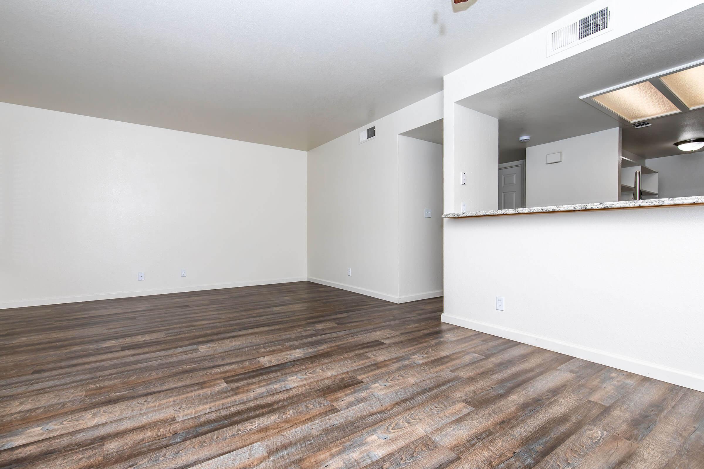 A spacious, empty room with light-colored walls and dark wooden flooring. A kitchen area is visible in the background with a bar counter. There are no furnishings, creating a minimalist and airy atmosphere. Natural light brightens the space, highlighting the clean and modern layout.