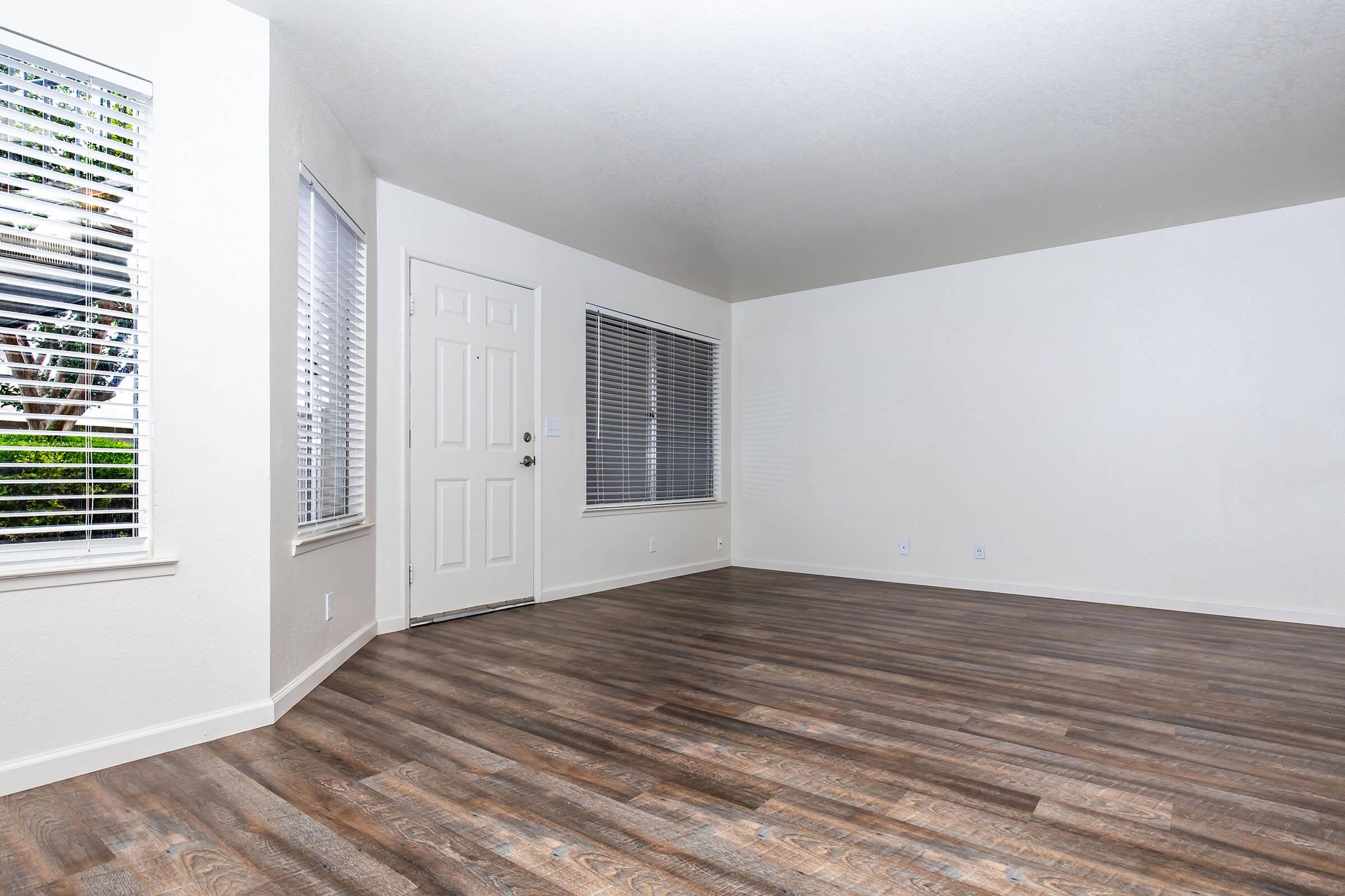 A spacious, empty living room with natural light coming from windows. The walls are painted in a light color, and the flooring is a warm brown laminate. There is a closed door leading outside, and the windows have white blinds partially drawn. The room is clean and ready for furniture.