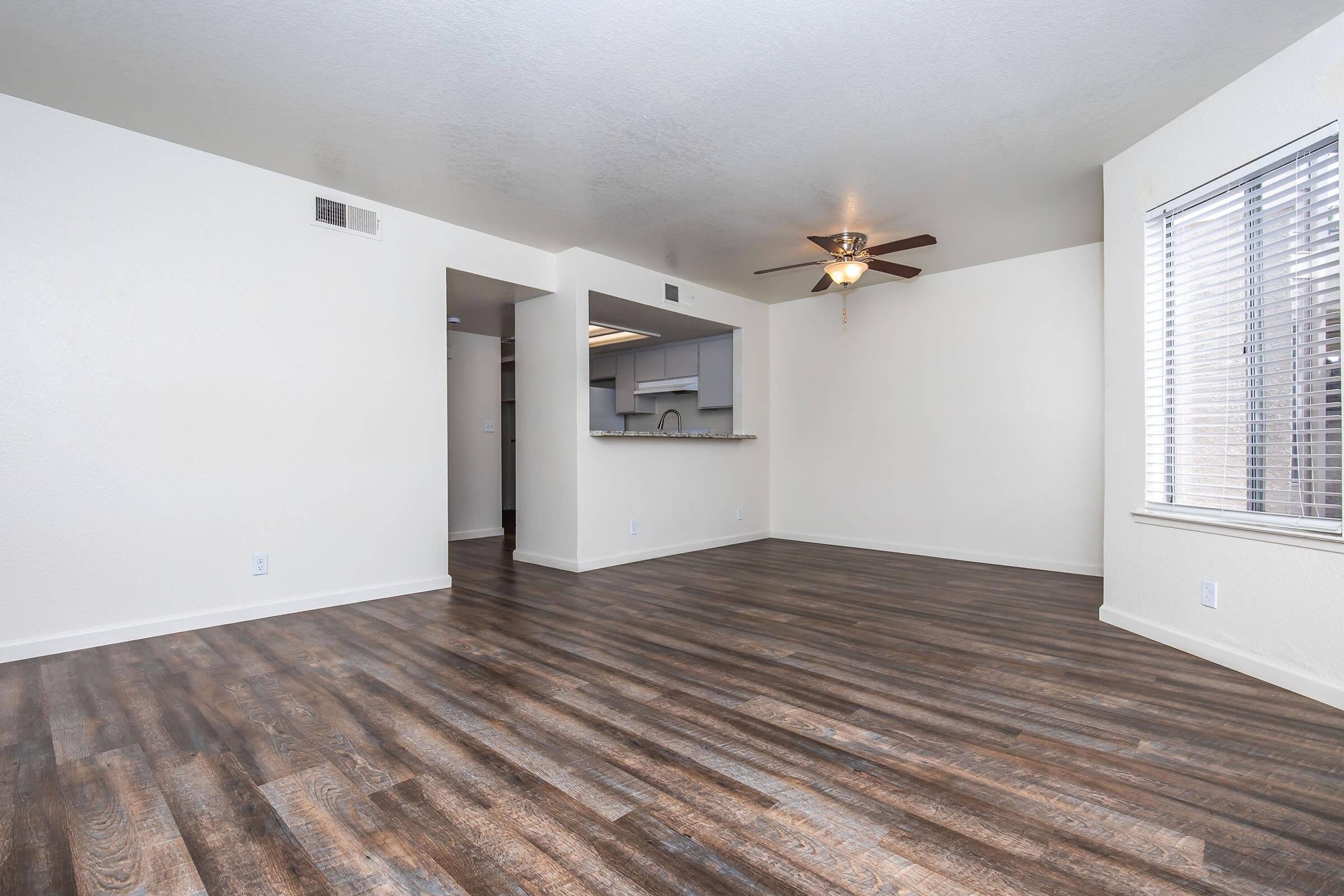A spacious, empty living room with light-colored walls and a ceiling fan. The flooring is dark wood-like laminate. To the left, there's an open kitchen area with a small countertop. A window with blinds is on the right side, allowing natural light into the room. The overall atmosphere is bright and airy.