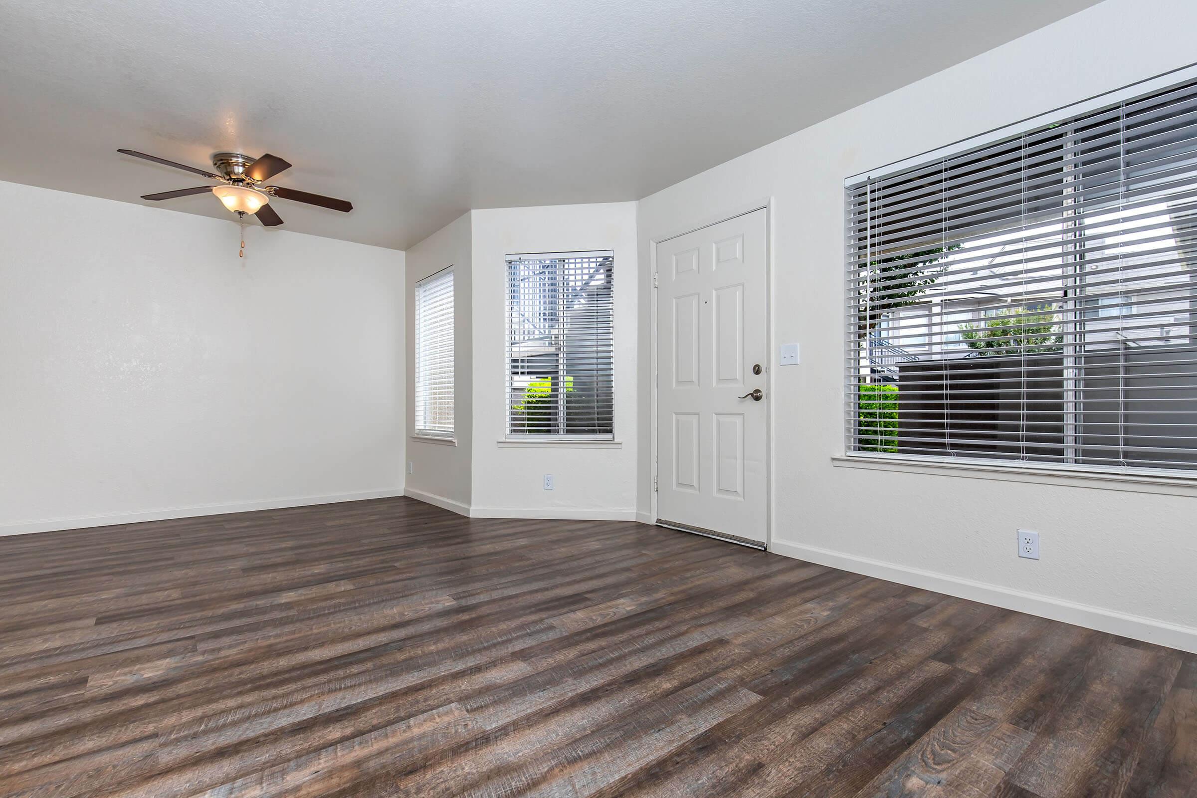 A spacious, empty living room with light-colored walls and dark wood-like flooring. There is a ceiling fan with light fixtures and windows with white blinds letting in natural light. A front door is visible, and the overall ambiance is bright and inviting, suitable for various interior designs.