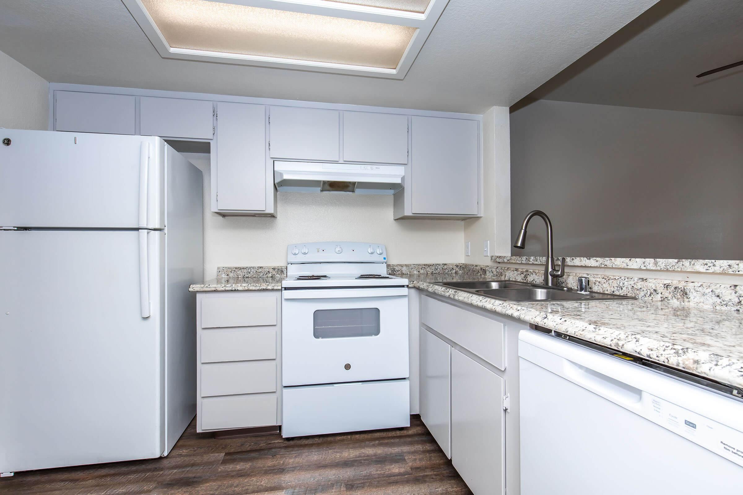 A modern kitchen featuring white cabinets, a white refrigerator, a white stove with an overhead microwave, and a large granite countertop. There is a stainless steel sink and a dishwasher under the counter. The floor has a wood-like appearance, and the space is well-lit with natural light.