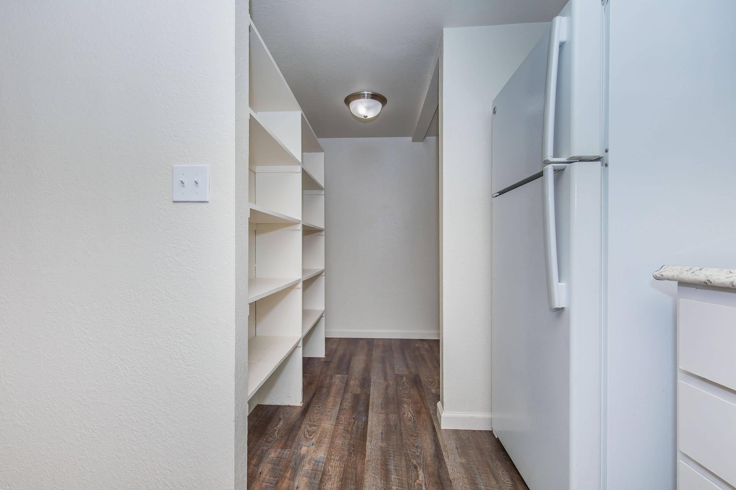 A narrow hallway features white shelves on the left and a refrigerator on the right. The walls are painted in light colors, and the flooring is wood-like, creating a bright and clean atmosphere. A ceiling light provides illumination in the space.