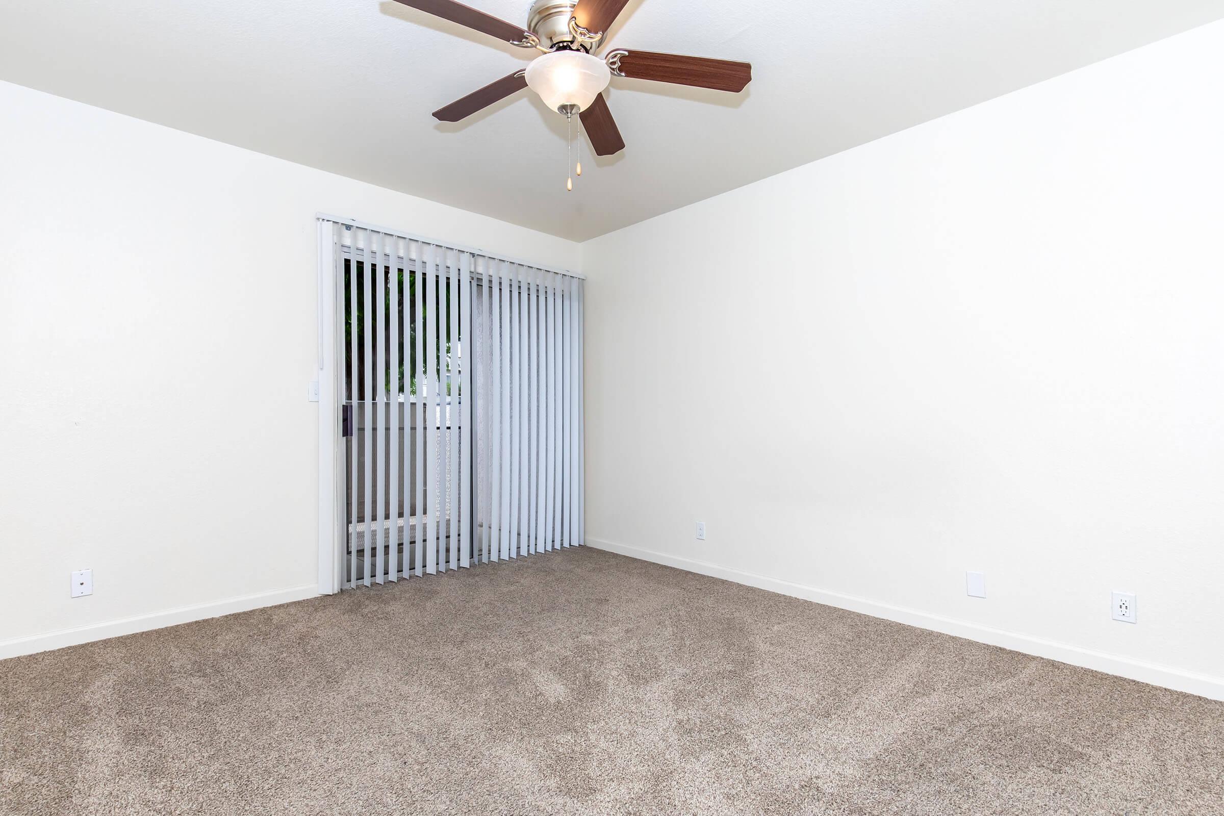 A spacious, empty room featuring beige carpet and light-colored walls. The ceiling includes a ceiling fan with wooden blades. A sliding glass door with vertical blinds leads to the outside. The overall ambiance is bright and airy, perfect for personalizing as a living space.