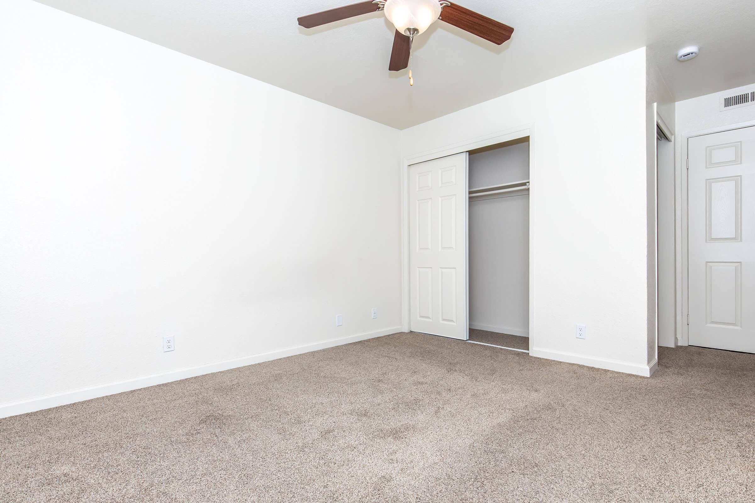 Empty room with light-colored walls and carpeted floor. A ceiling fan is mounted on the ceiling, and one corner has a closed double-door closet. The room appears spacious and well-lit, with a simple layout and no furniture present.