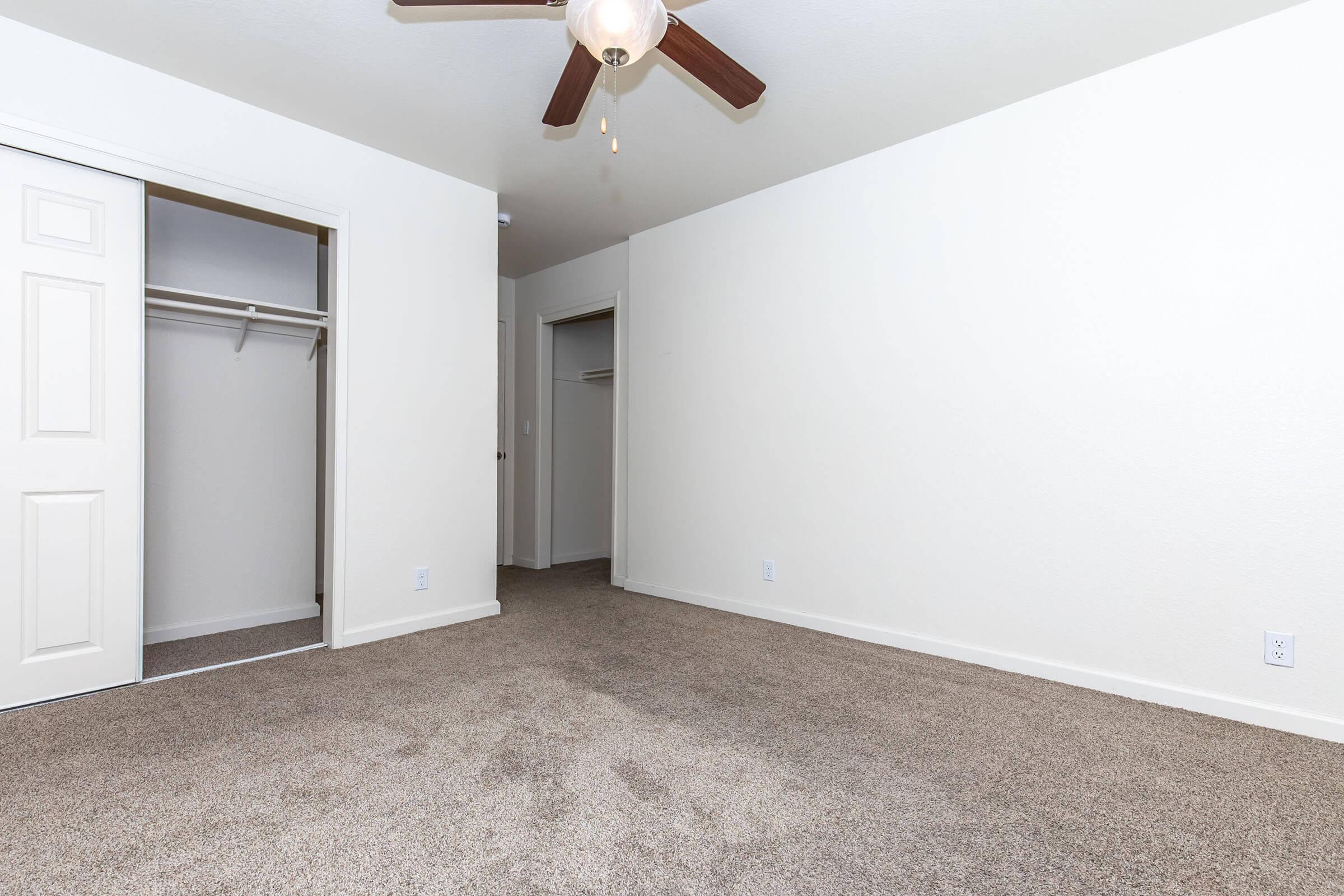 A spacious room with a ceiling fan, light-colored walls, and carpeted flooring. There are two open doorways leading to closets, with one visible and the other partially obscured. The room has a neutral color scheme and appears bright and inviting, suitable for various uses.