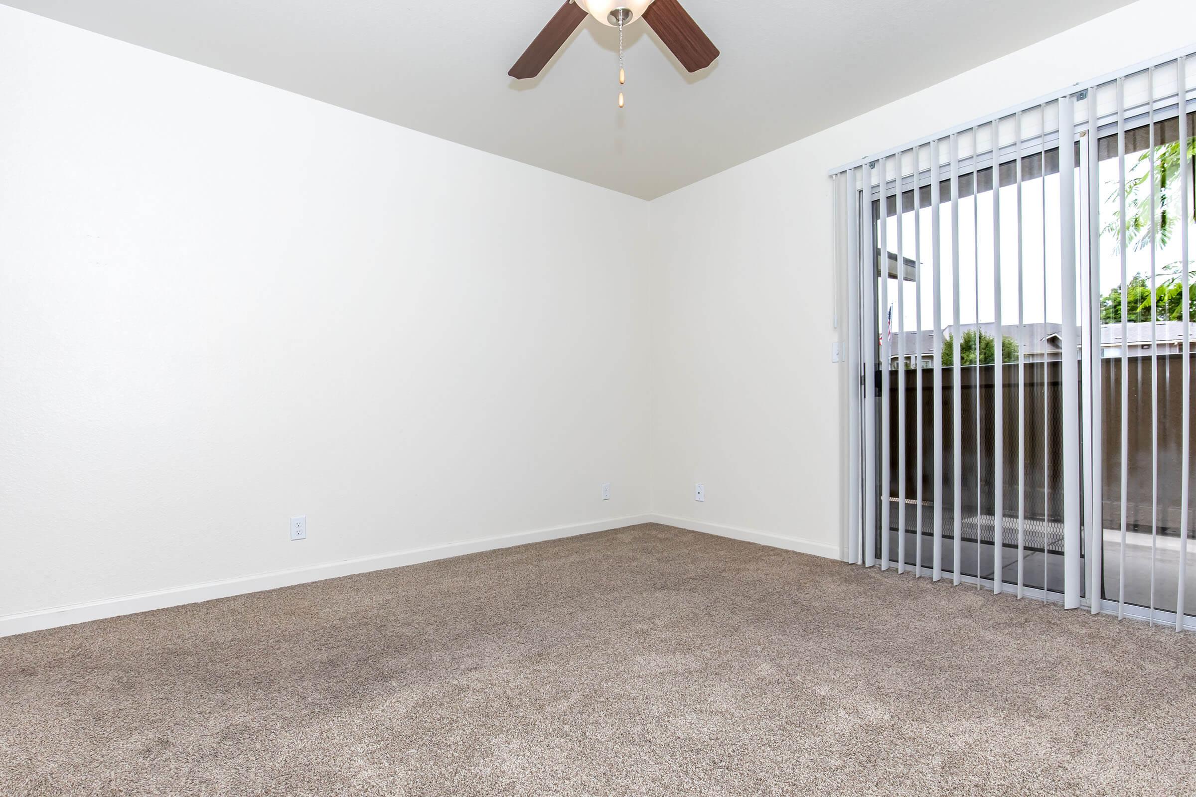 A spacious room with beige carpet and white walls, featuring a ceiling fan with wooden blades. There are vertical blinds covering a large window that leads to a patio area, letting in natural light. The room is empty, creating a blank canvas for personal design.