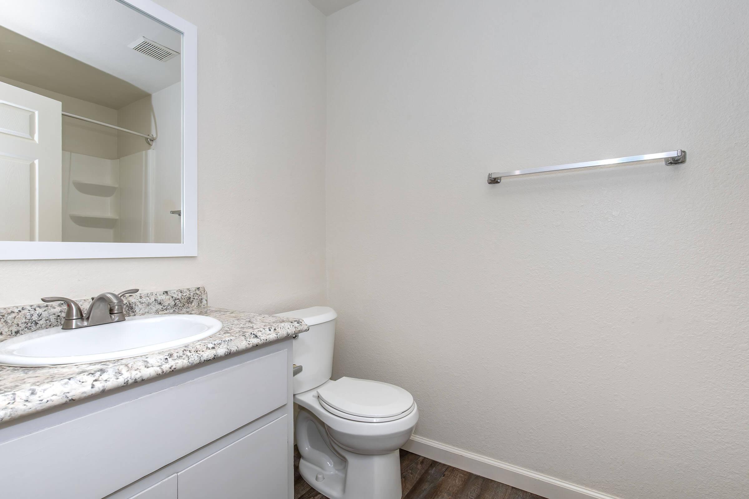 A clean and modern bathroom featuring a white countertop sink with a mirror above, a toilet, and a towel rack on the wall. The walls are a light color, and the flooring has a wood-like appearance, contributing to a fresh and simple design.