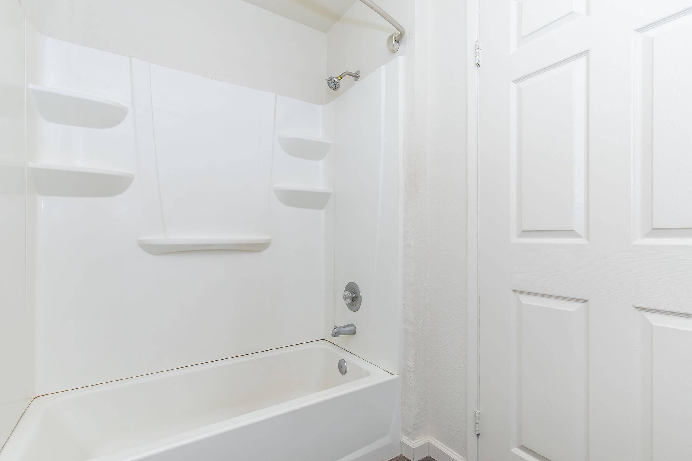 A clean, modern bathroom featuring a white bathtub with a built-in shower. The tub has a wall-mounted showerhead and a sliding shower curtain rod. The walls are painted a light color, and there are two built-in shelves for storage. A white door is visible in the background.