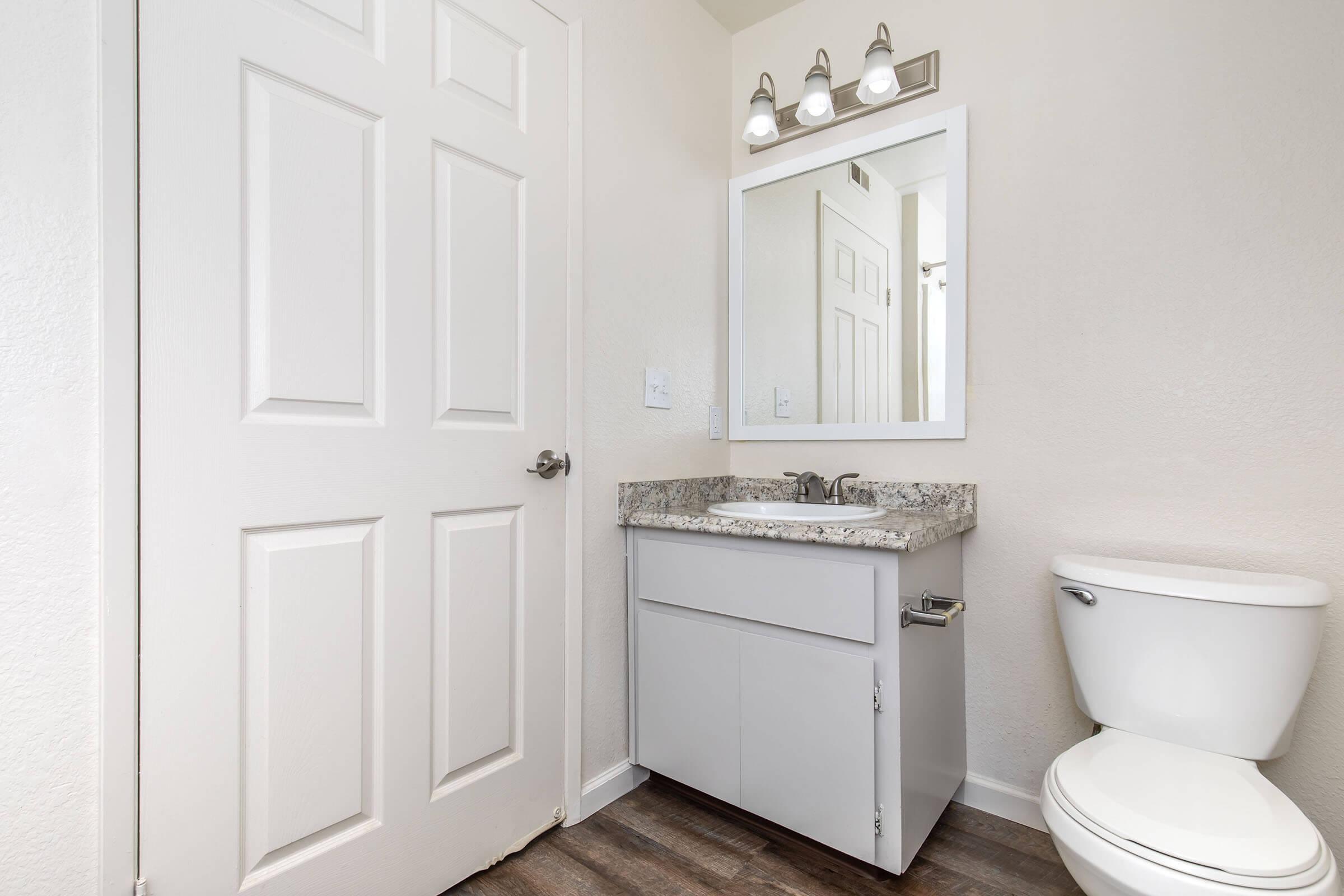 A small bathroom featuring a white vanity with a granite countertop and a sink, a mirror mounted above the vanity, and a toilet to the right. There is a closed white door on the left, and the floor is covered with wooden laminate. The walls are painted a light color, enhancing the brightness of the space.