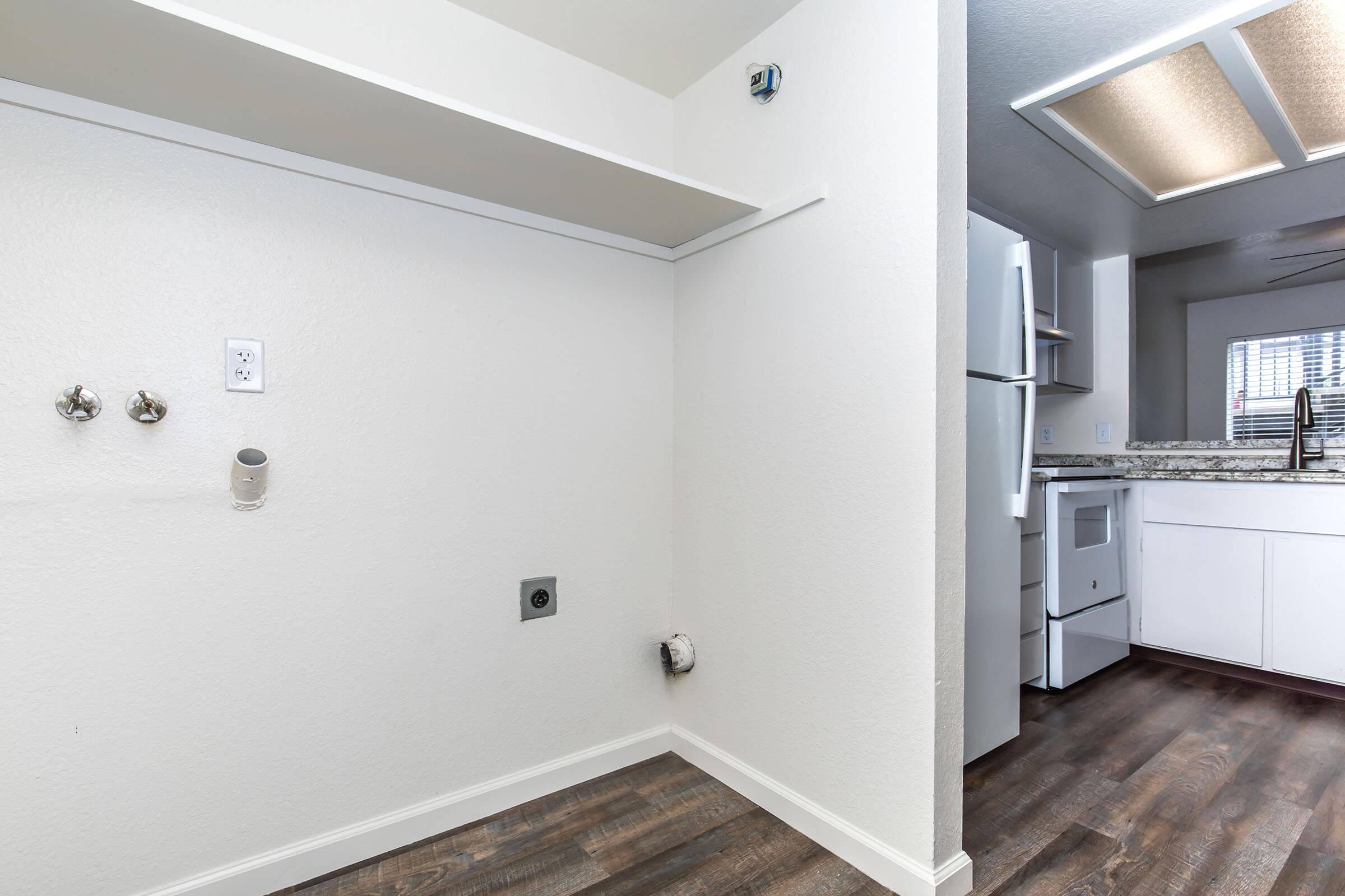 A well-lit corner of a modern kitchen featuring white walls and hardwood floors. The area includes exposed plumbing connections on the left, with a small shelf above. In the background, a refrigerator and a part of the kitchen counter and appliances are visible, emphasizing a clean and functional design.