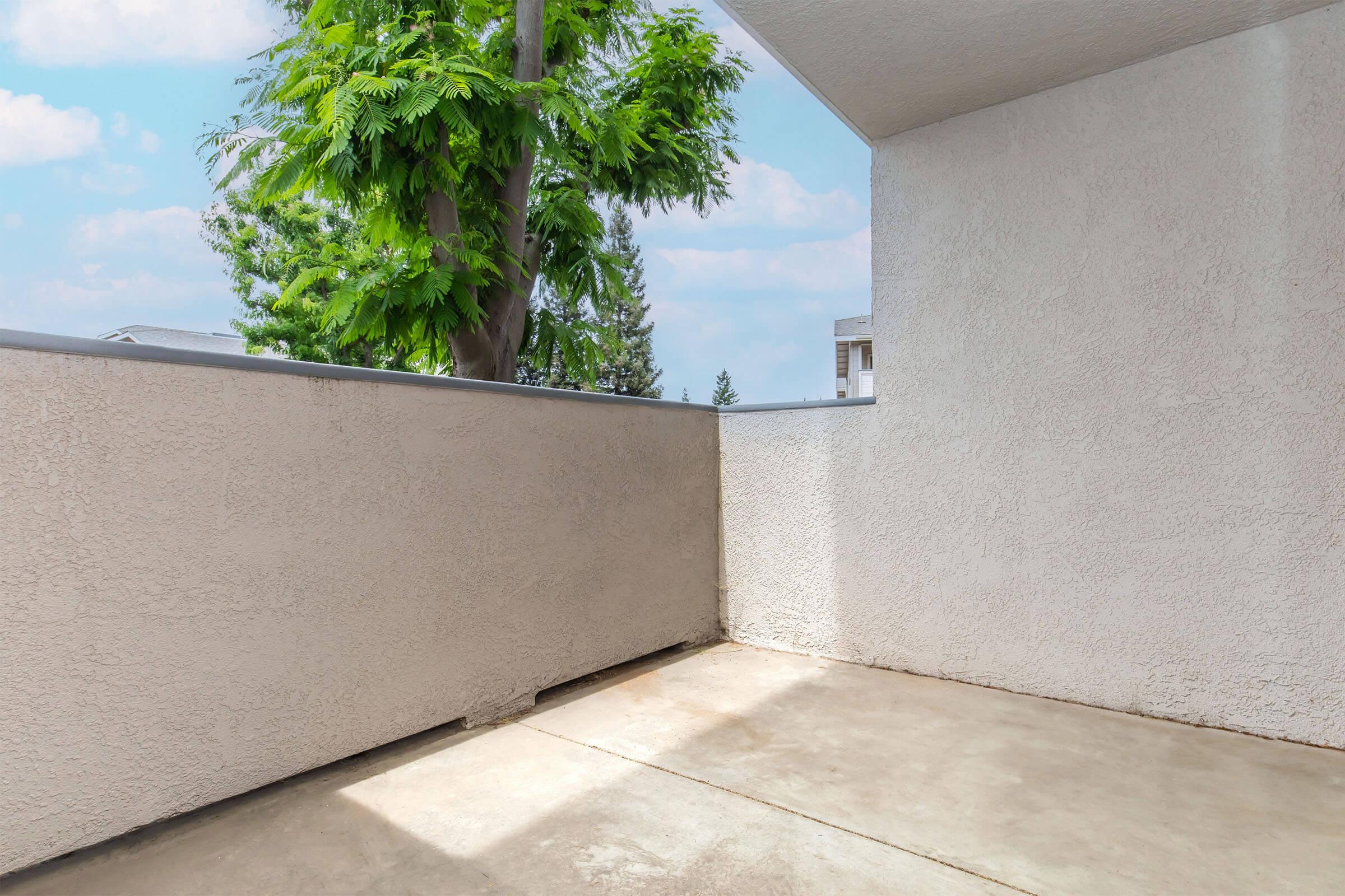 A clean, bare balcony with a concrete floor. A textured white wall is visible on one side, and a lush green tree can be seen outside. The sky is blue with a few clouds in the background, creating a bright and airy atmosphere.