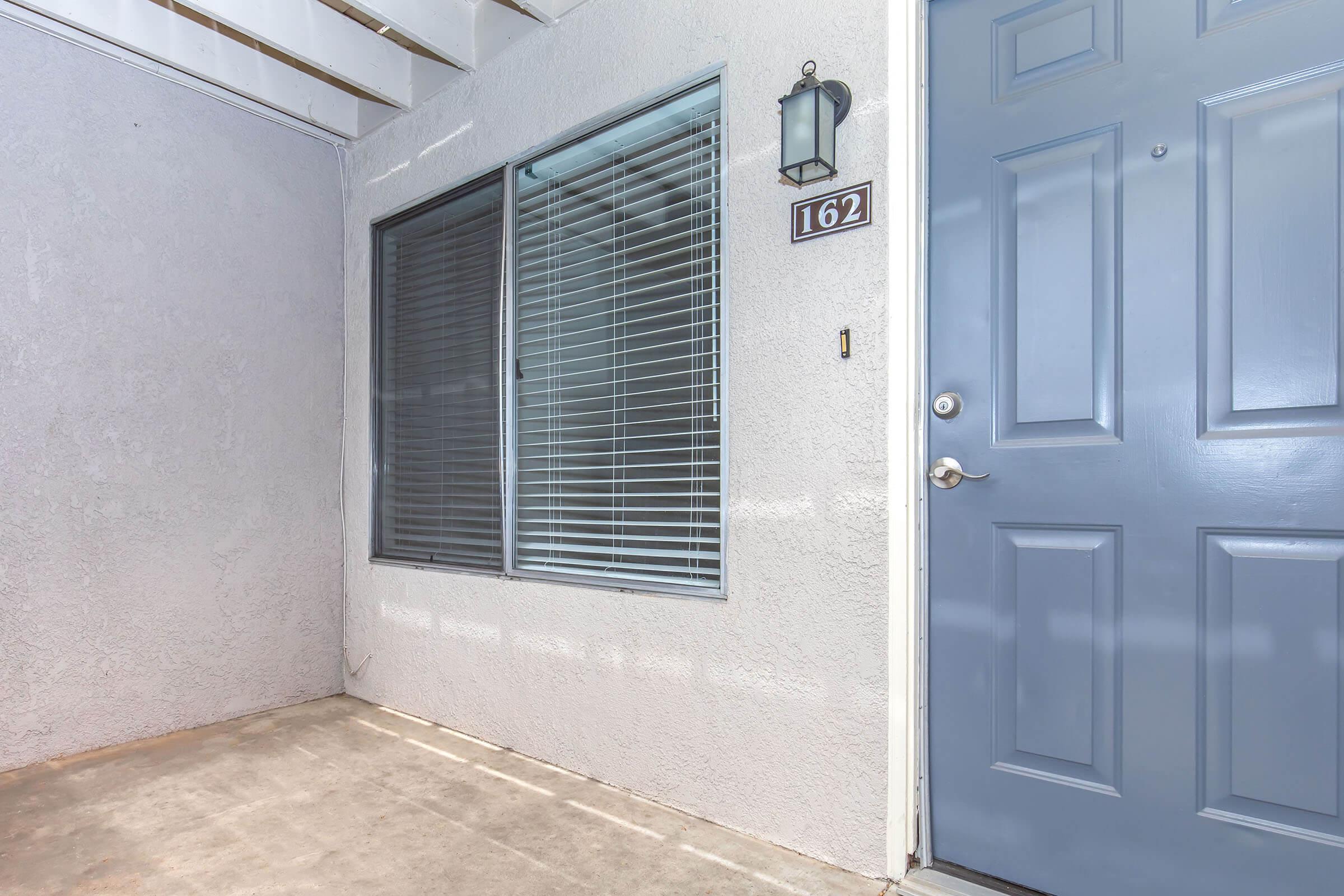 A view of the entrance to a building, featuring a blue door with the number 162 attached. There is a window beside the door with horizontal blinds, and the wall is a light gray color. The floor appears to be concrete and the area is sheltered by an overhang.