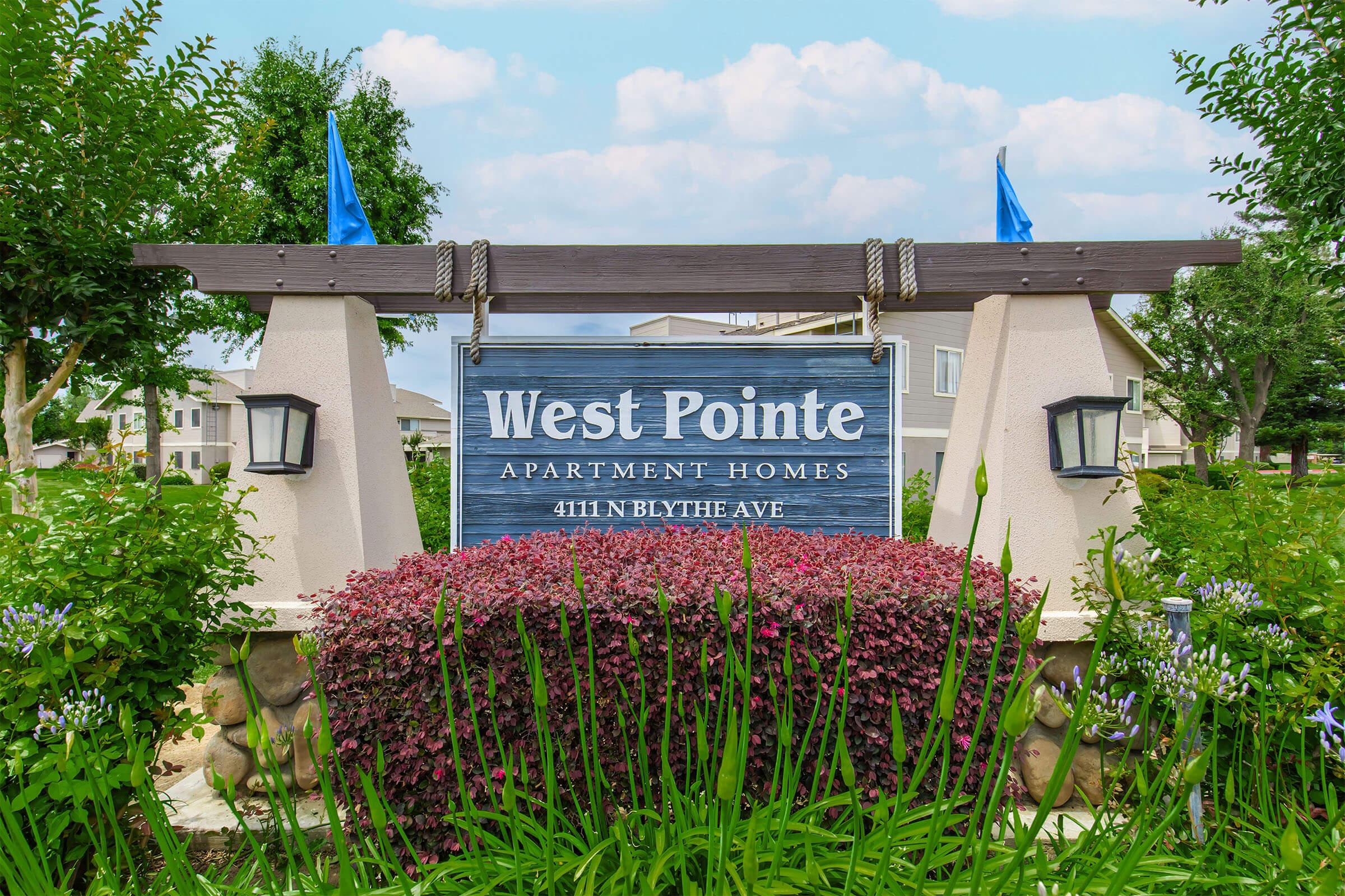 Sign for West Pointe Apartment Homes, located at 4111 N Blythe Ave. The sign features decorative elements like blue flags and lanterns, surrounded by colorful shrubs and green plants under a partly cloudy sky.
