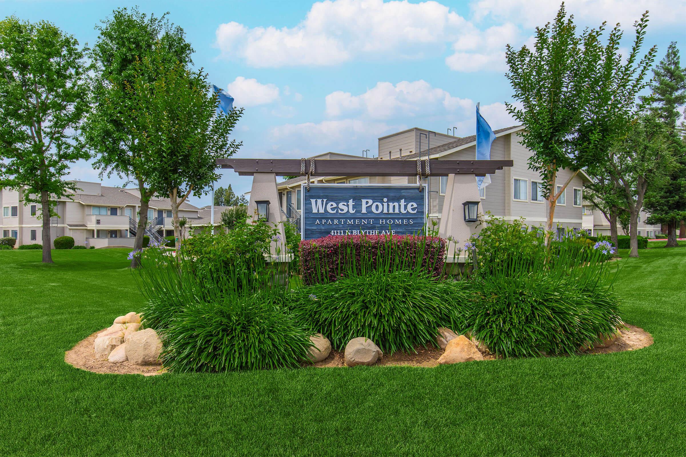 A sign for West Pointe Apartment Homes situated in a green lawn surrounded by shrubs and rocks. The background features residential buildings and a cloudy blue sky.