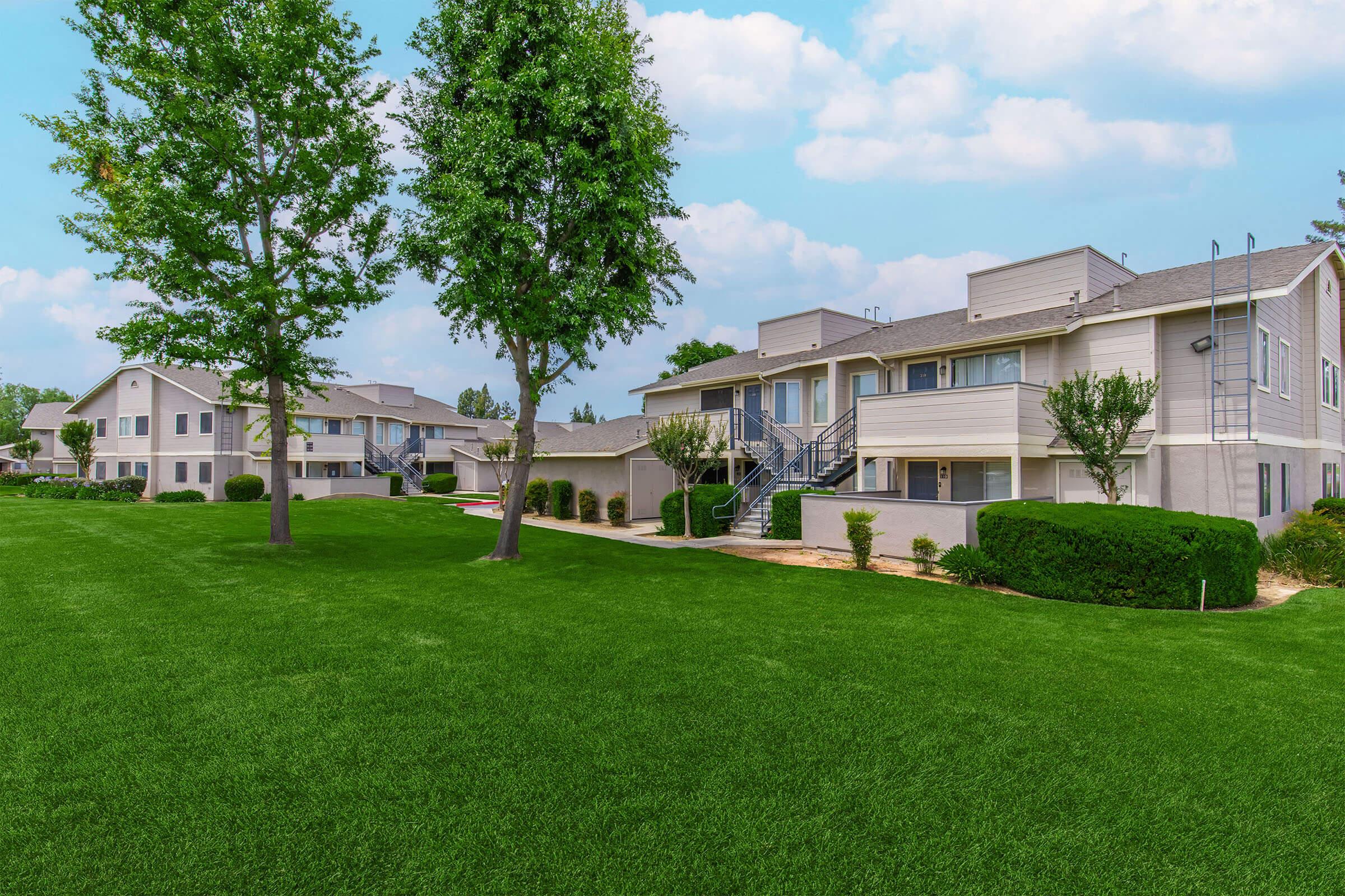 A residential area featuring two buildings with light-colored exteriors, surrounded by well-maintained green lawns and trees. The sky is partly cloudy, enhancing the inviting atmosphere of the neighborhood.