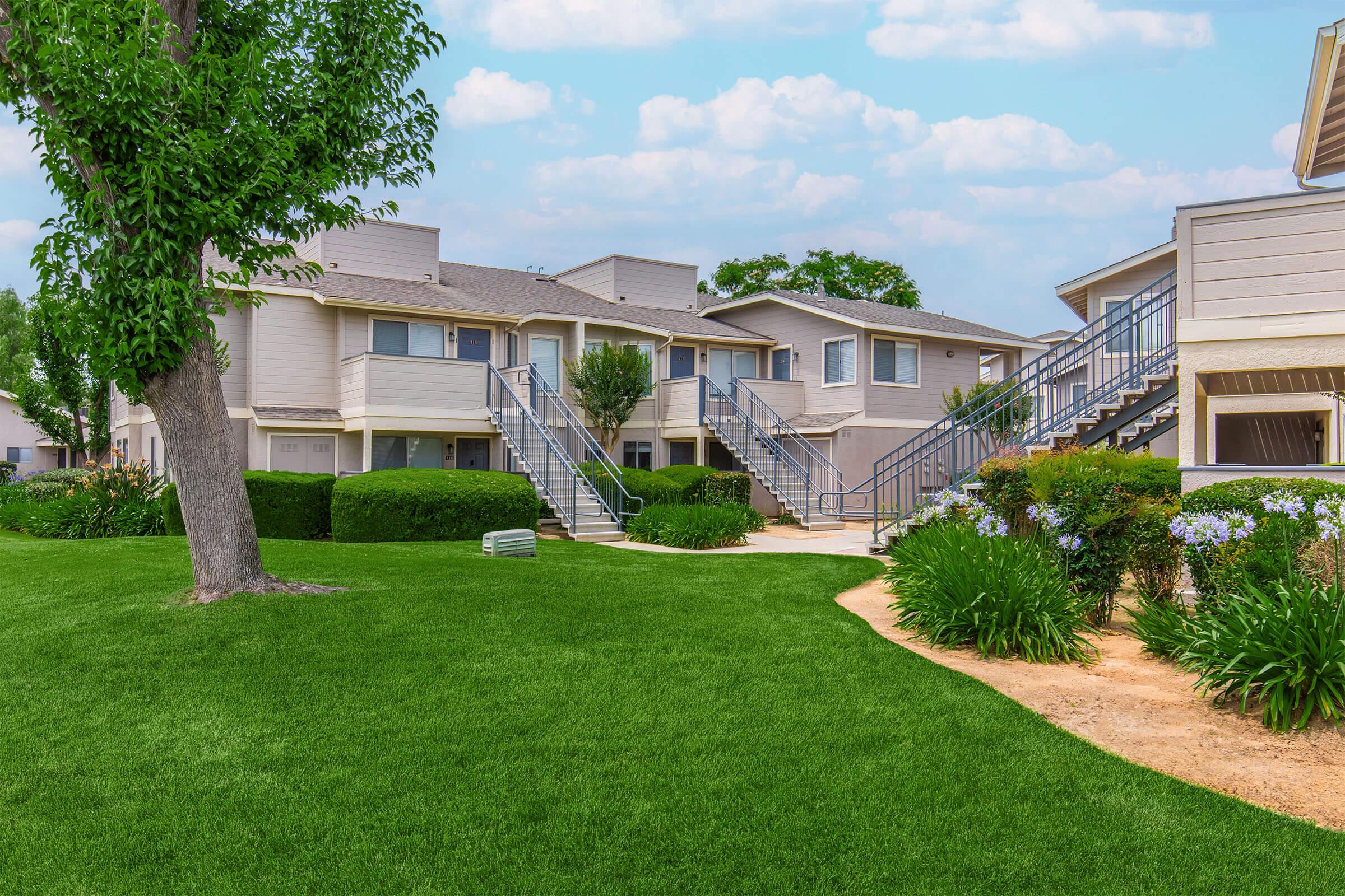 Lush green lawn in front of two-story apartment buildings with stairs leading to individual units. Trees and shrubs are present, contributing to a well-maintained landscape under a partly cloudy sky.
