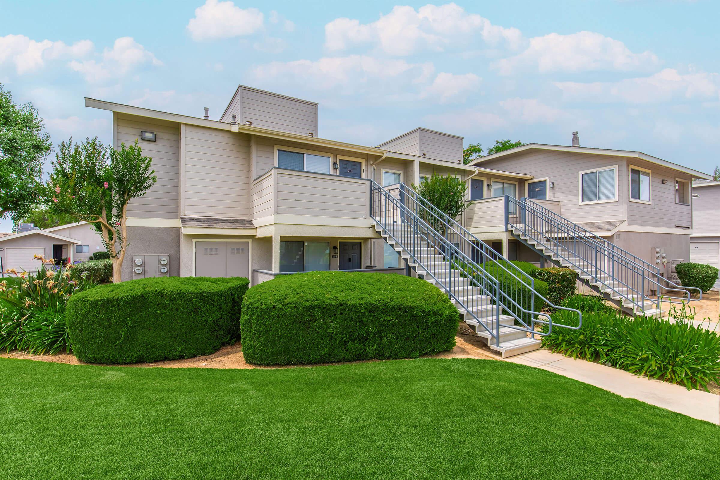 Two-story residential building with a modern design, featuring a light-colored exterior and multiple balconies. The building is surrounded by neatly trimmed bushes and green grass, with concrete pathways leading to the entrances. A clear blue sky is visible in the background.