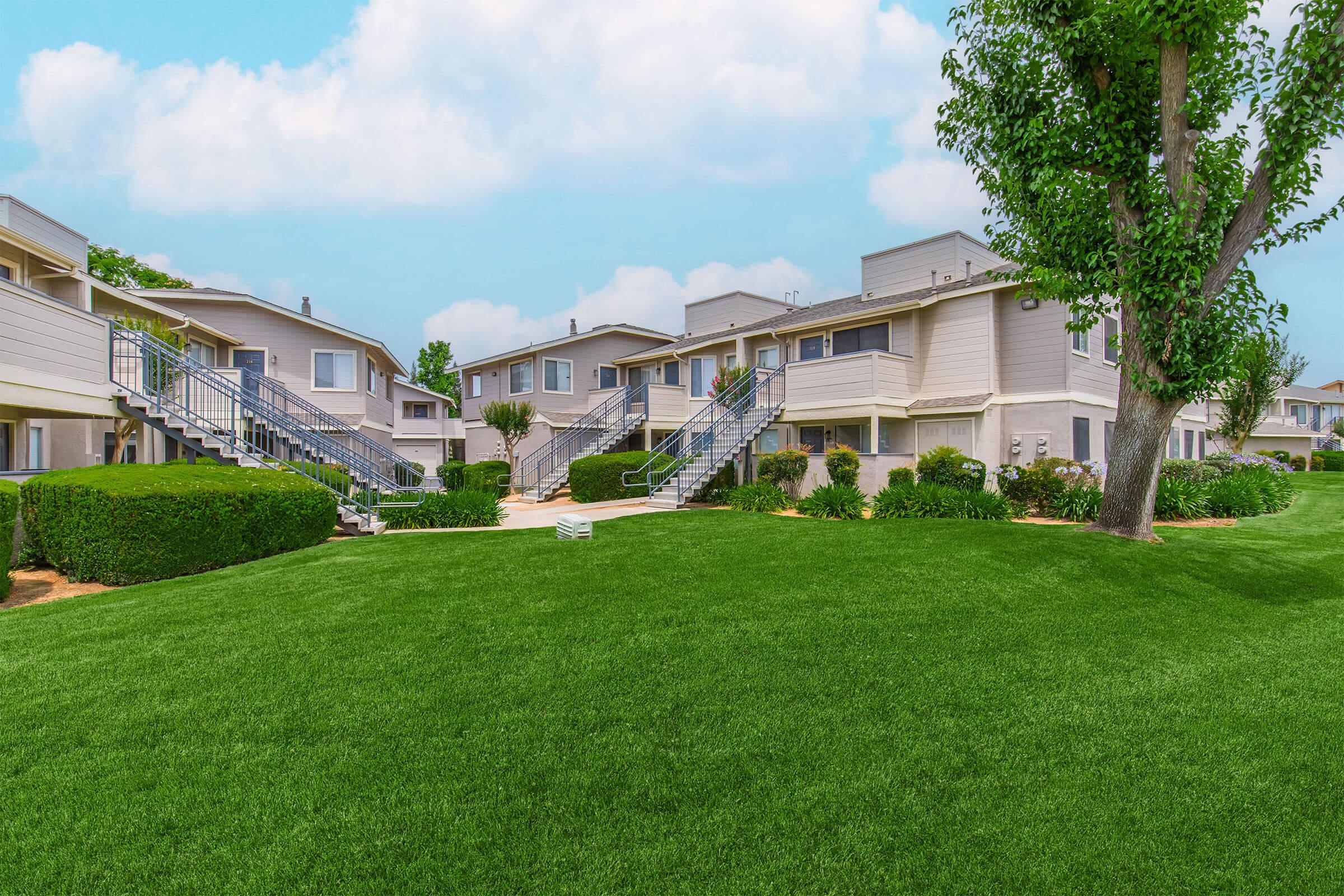 A well-maintained apartment complex with two-story buildings surrounded by lush green lawns and neatly trimmed bushes. There are trees providing shade, and clear blue skies in the background. The area appears inviting and landscaped for tenant enjoyment.