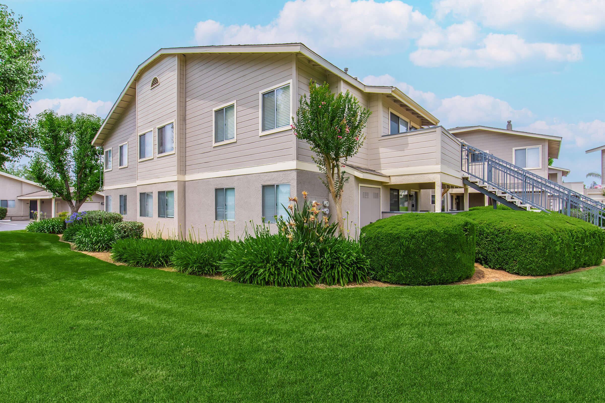 A well-maintained two-story apartment building surrounded by lush green grass and landscaped shrubs. The exterior features light-colored siding and a balcony. The scene is set against a clear blue sky with fluffy clouds, creating a peaceful and inviting atmosphere.
