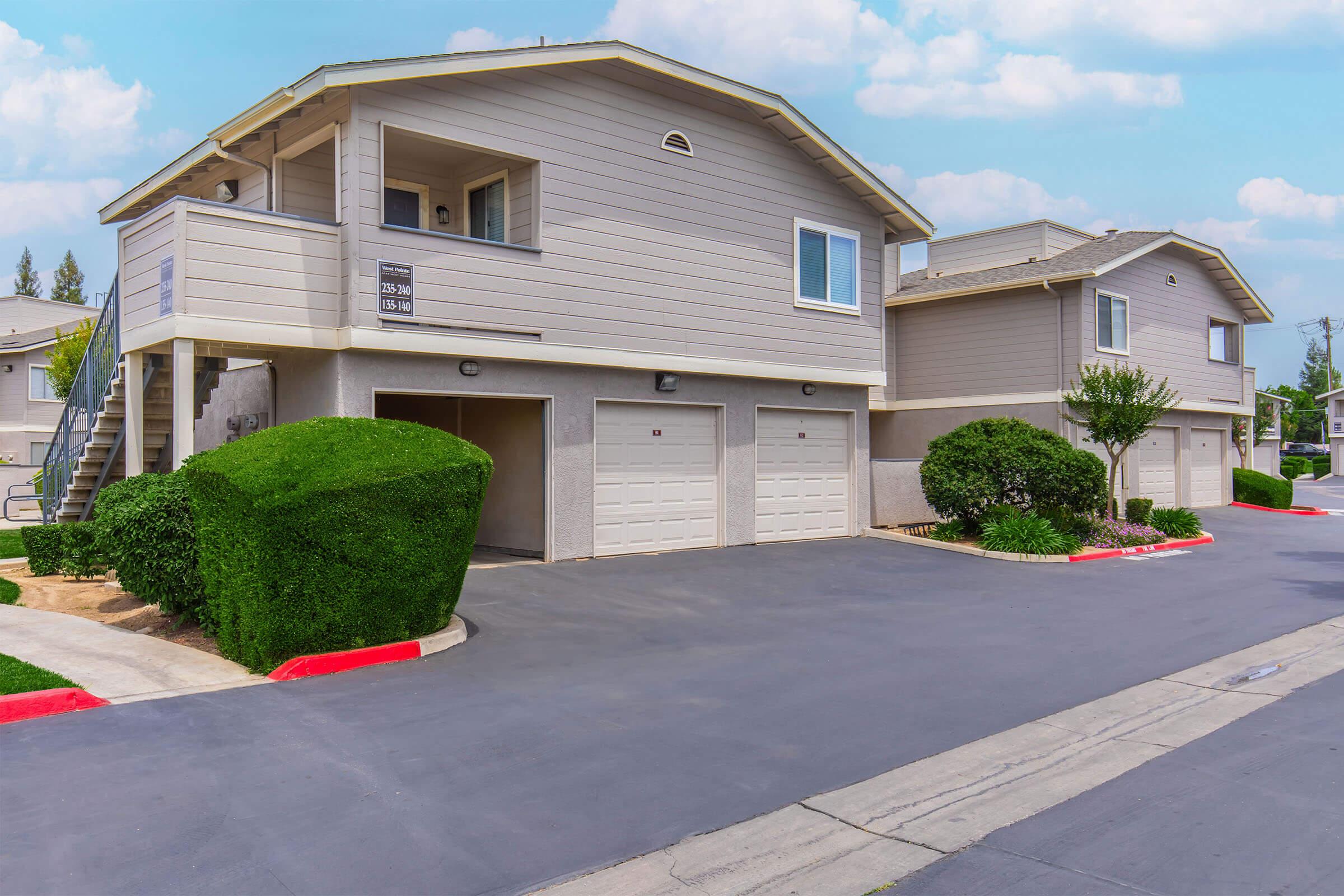 Two multi-story residential buildings with garages at the ground level. The buildings feature light gray siding and large windows. Well-maintained green shrubs are planted along the driveways, which lead to the garages. The scene is set against a clear blue sky with a few clouds.