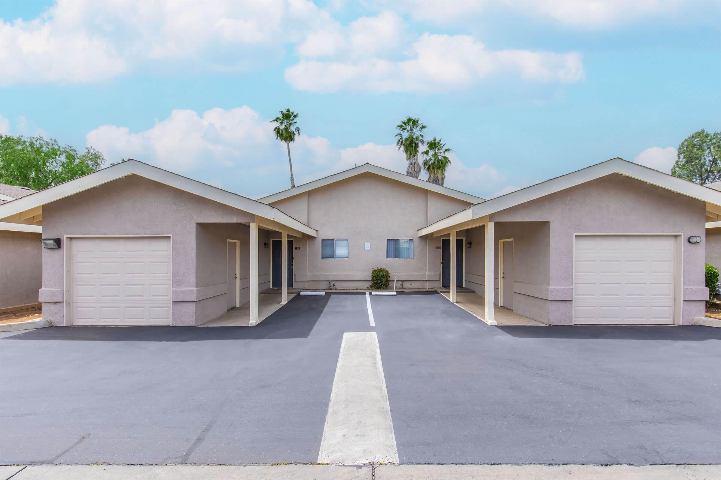Two identical single-story buildings with light-colored exteriors, each featuring a garage door. In between, there is a paved driveway leading to a central area. Palm trees are visible in the background under a partly cloudy blue sky. The setting appears serene and suburban.