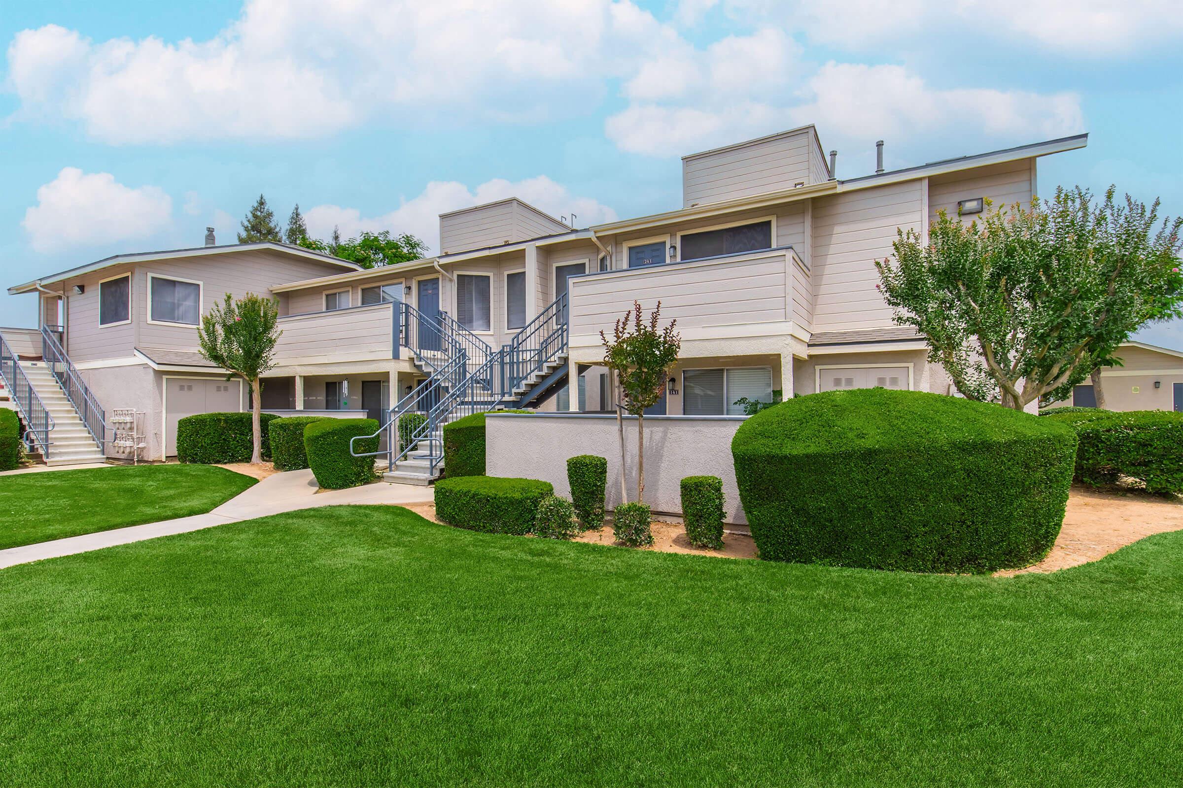 Exterior view of a residential apartment complex featuring two-story buildings with stairs. The well-maintained landscape includes manicured bushes and a green lawn. The sky is partly cloudy, and there are walkways connecting the units.