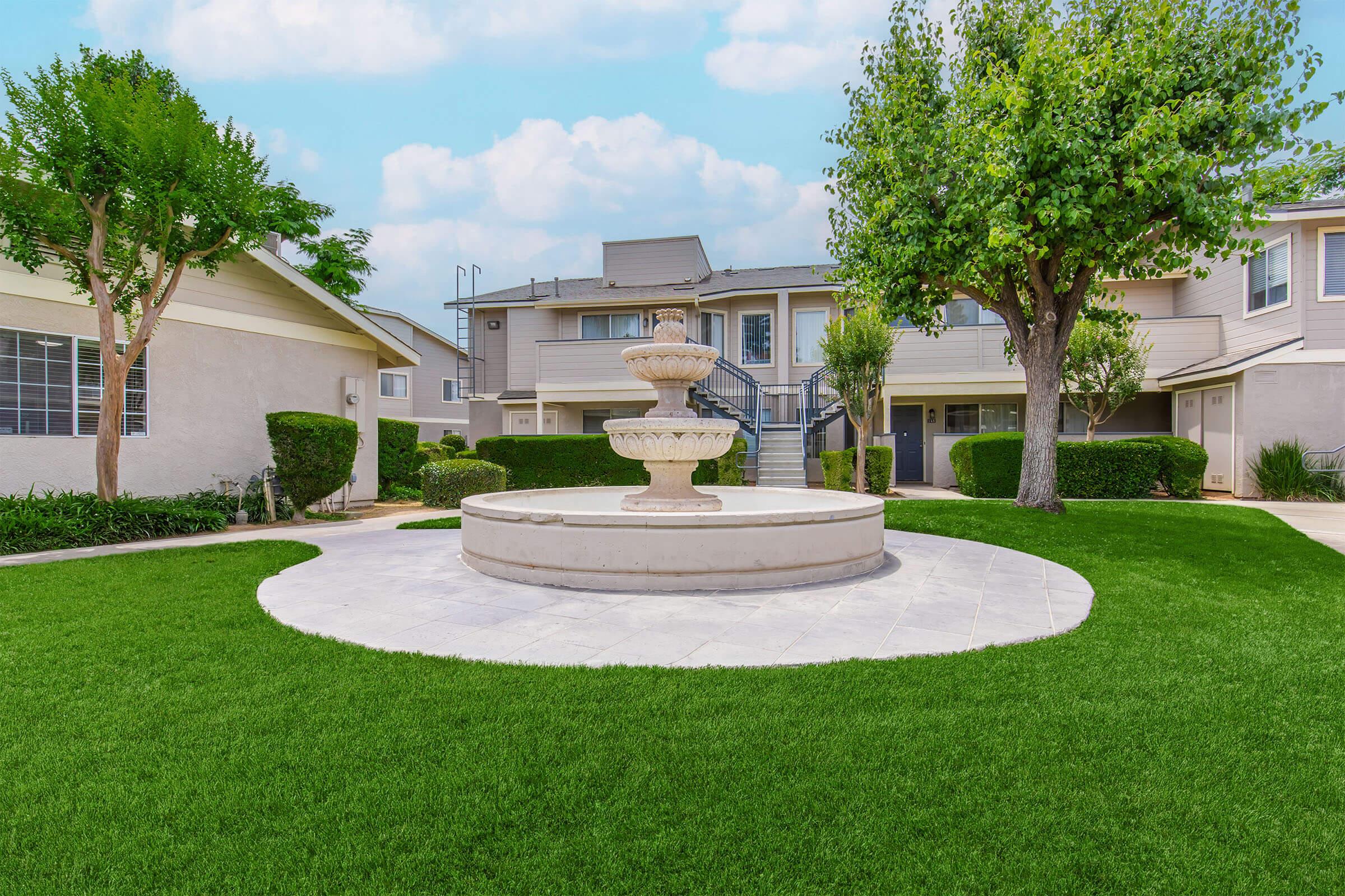 A landscaped courtyard featuring a decorative fountain at the center, surrounded by neatly trimmed grass and shrubbery. In the background, there are two multi-story buildings with balconies and trees lining the area, creating a serene outdoor space.