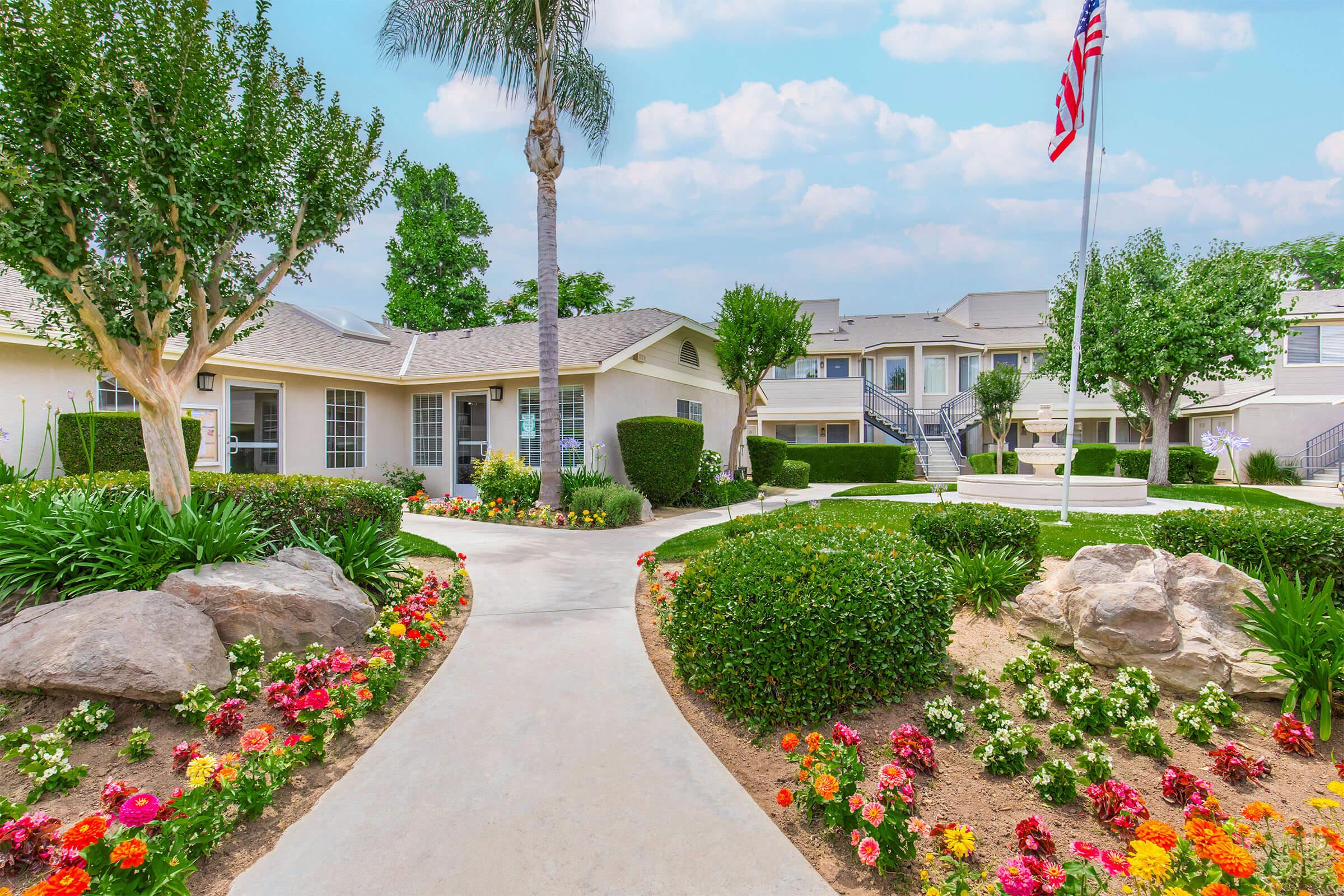 A well-maintained landscape featuring a pathway lined with colorful flowers. On the left, there are small trees and shrubs, while a building can be seen in the background. In the foreground, a palm tree adds tropical flair. The scene is bright and inviting, with a clear blue sky above.