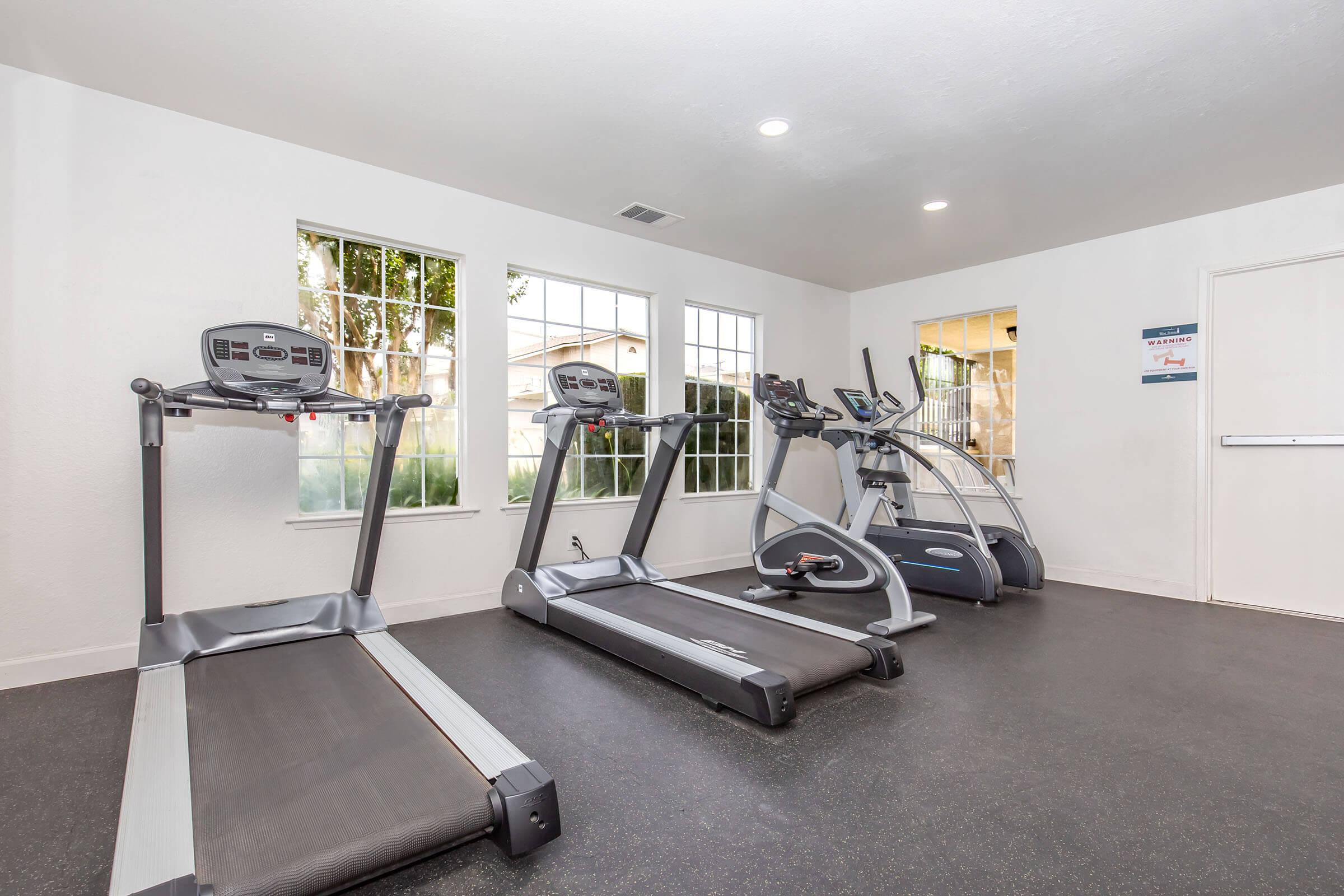 A bright and clean gym space featuring three pieces of exercise equipment: two treadmills and an elliptical machine. Large windows let in natural light, creating an open and inviting atmosphere. The floor is dark, and there are minimal decorations, focusing on the workout equipment.