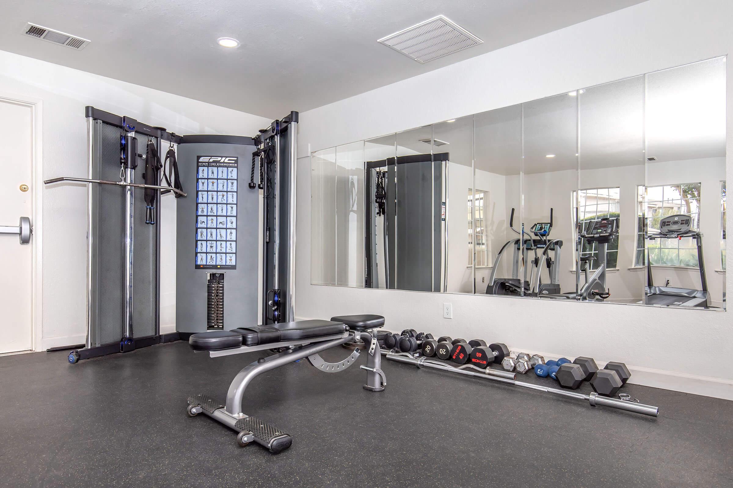 A modern home gym featuring a multi-function workout machine, a weight bench, and a variety of dumbbells arranged on the floor. A large mirror on the wall reflects the gym equipment, creating an open and spacious feel in the workout area. Natural light streams in from a window.