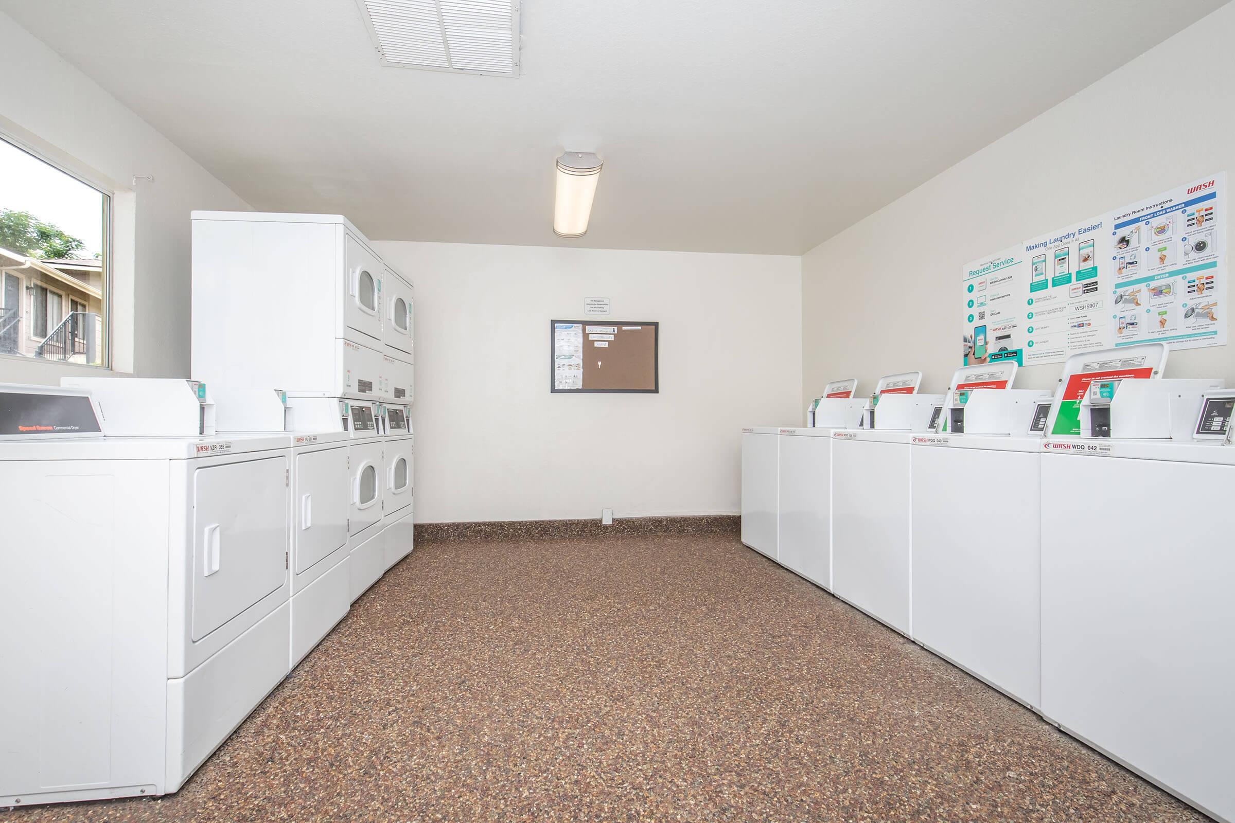 A clean, well-lit laundry room featuring several white washers and dryers lined against the walls. There is a bulletin board and instructional posters on the wall, with a window letting in natural light. The floor is covered in smooth, dark speckled tiles.
