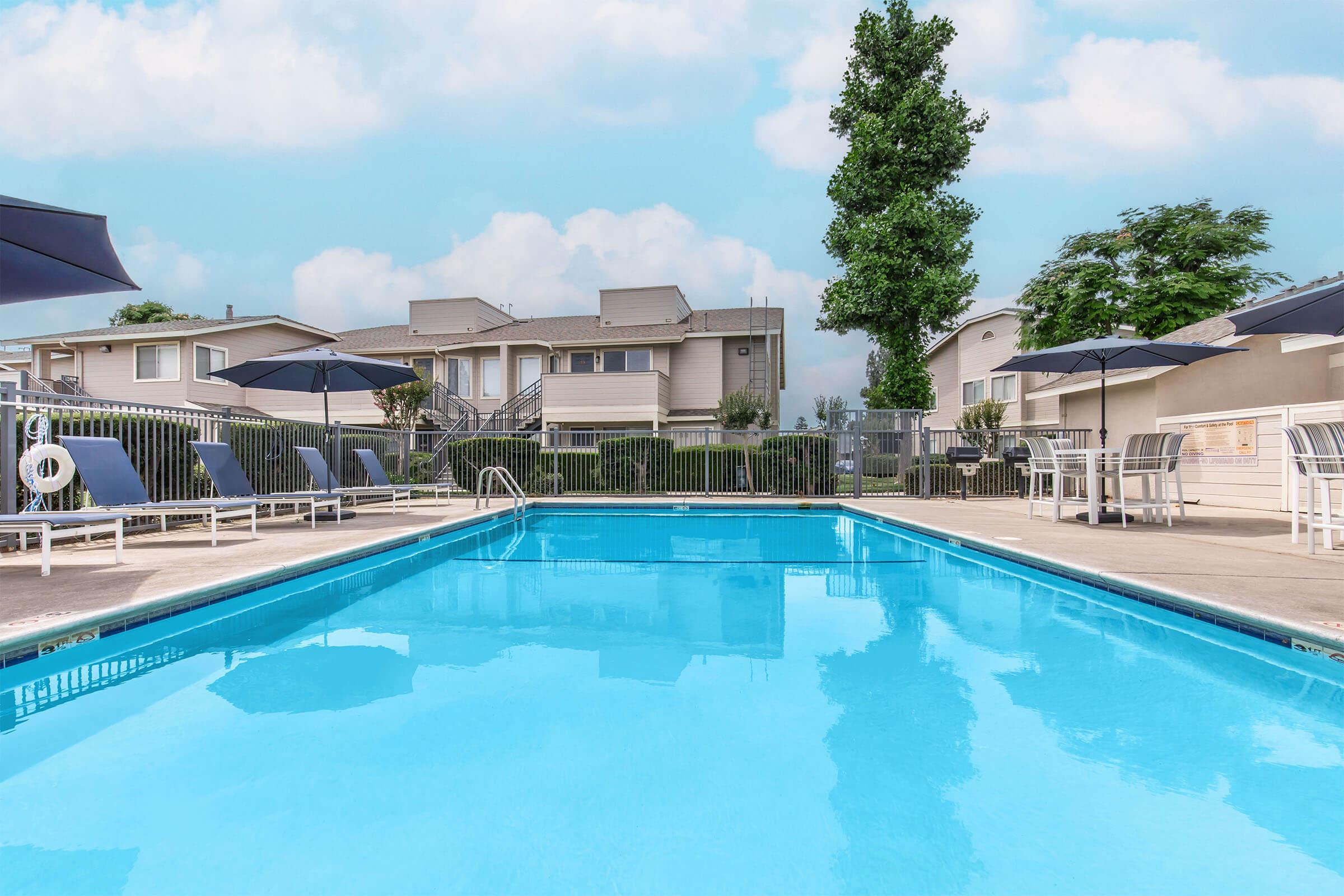 A clear blue swimming pool surrounded by lounge chairs and umbrellas, with a backdrop of apartment buildings and green trees under a bright sky. The scene creates a relaxing atmosphere for enjoying outdoor leisure activities.