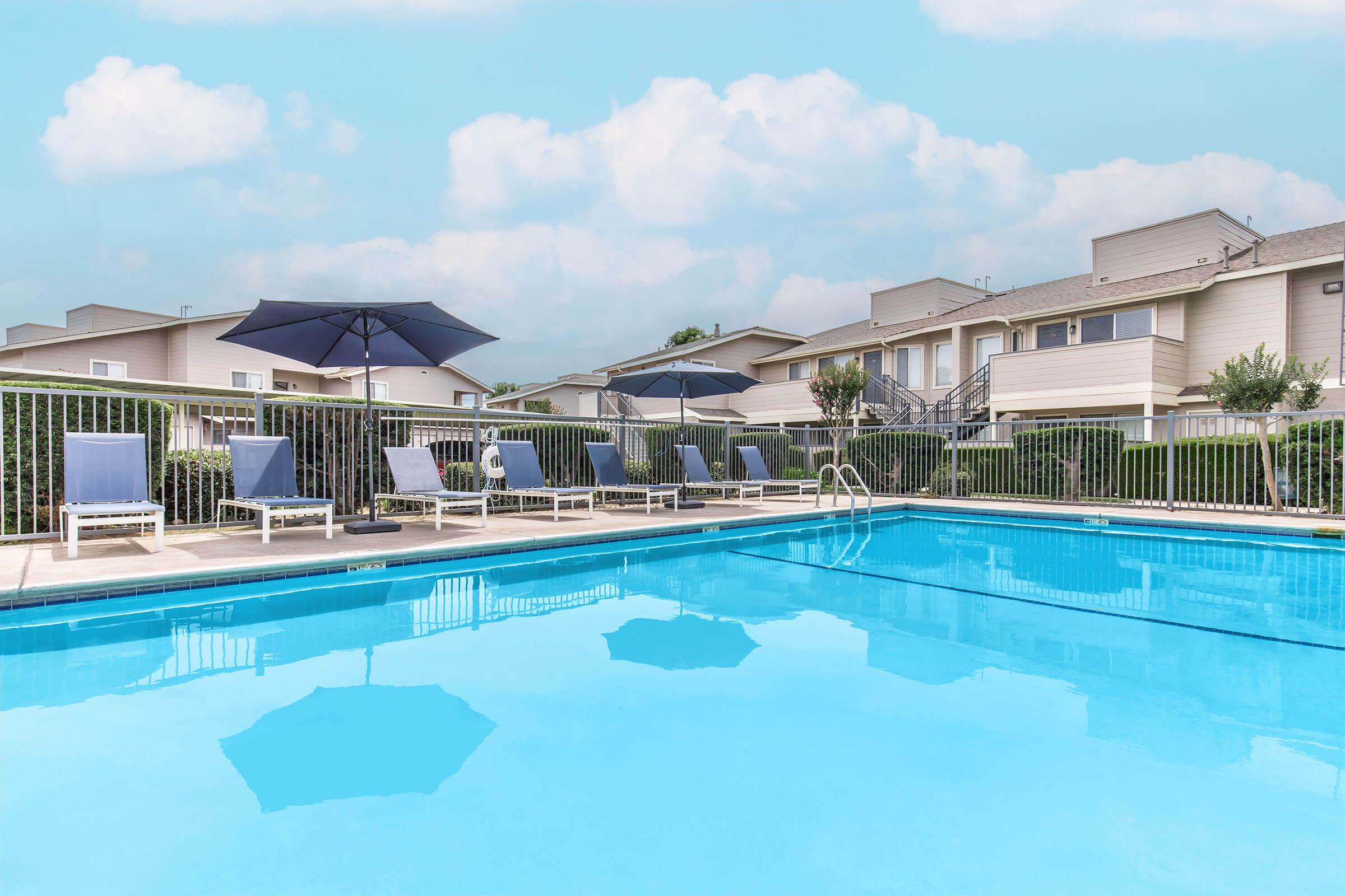 A clear blue swimming pool surrounded by lounge chairs and umbrellas, with well-maintained townhouses in the background under a bright sky with scattered clouds. The scene conveys a relaxing outdoor atmosphere ideal for leisure and enjoyment.