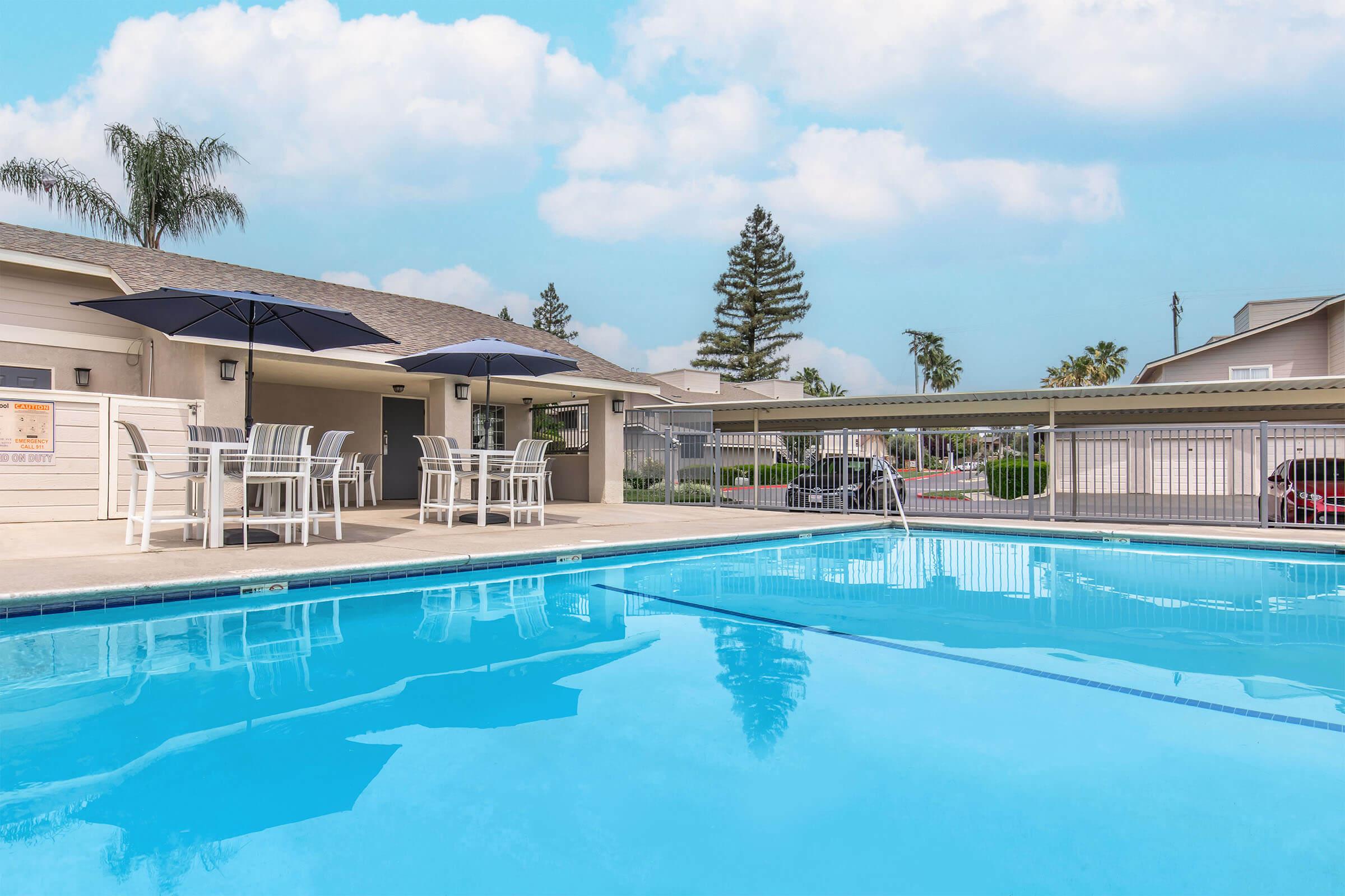 A clear swimming pool surrounded by a patio with tables and umbrellas. In the background, houses and palm trees are visible under a blue sky with sparse clouds. The area appears inviting and well-maintained, ideal for relaxation or recreation.