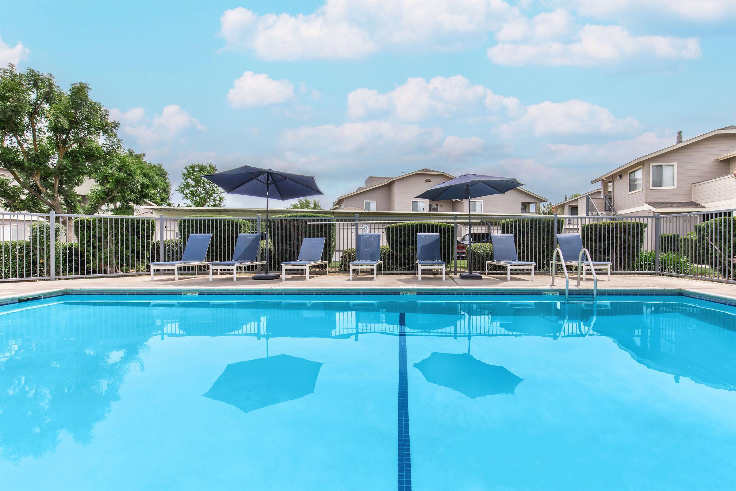 A clear swimming pool surrounded by lounge chairs and umbrellas. The pool reflects the blue sky and fluffy clouds above. In the background, there are residential buildings and green bushes, creating a relaxing outdoor atmosphere.