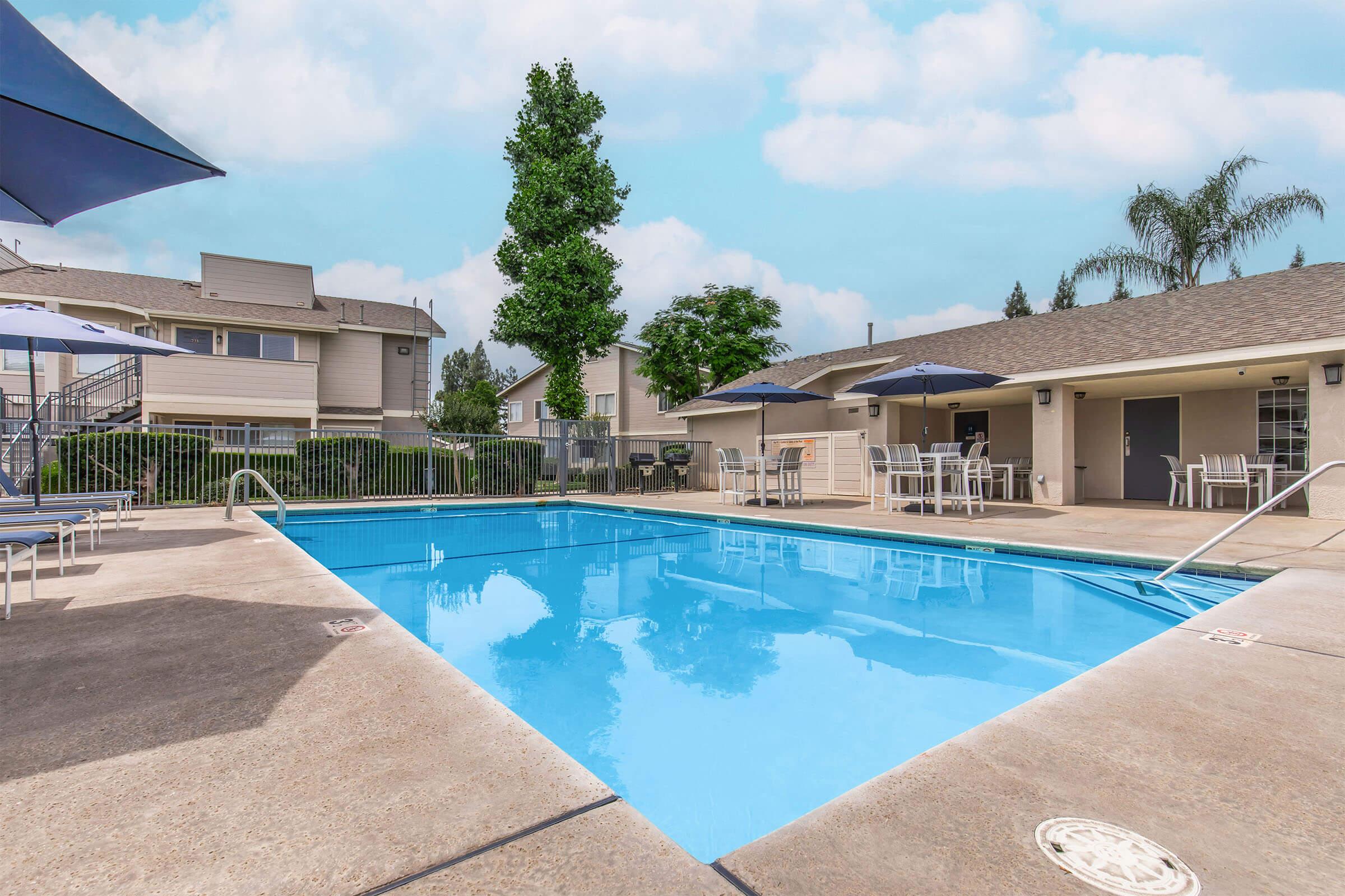 A view of a swimming pool surrounded by a concrete patio and lounge chairs. There are umbrellas for shade, and nearby are several residential buildings with greenery in the background. The sky is light blue with a few clouds, creating a bright and inviting atmosphere.
