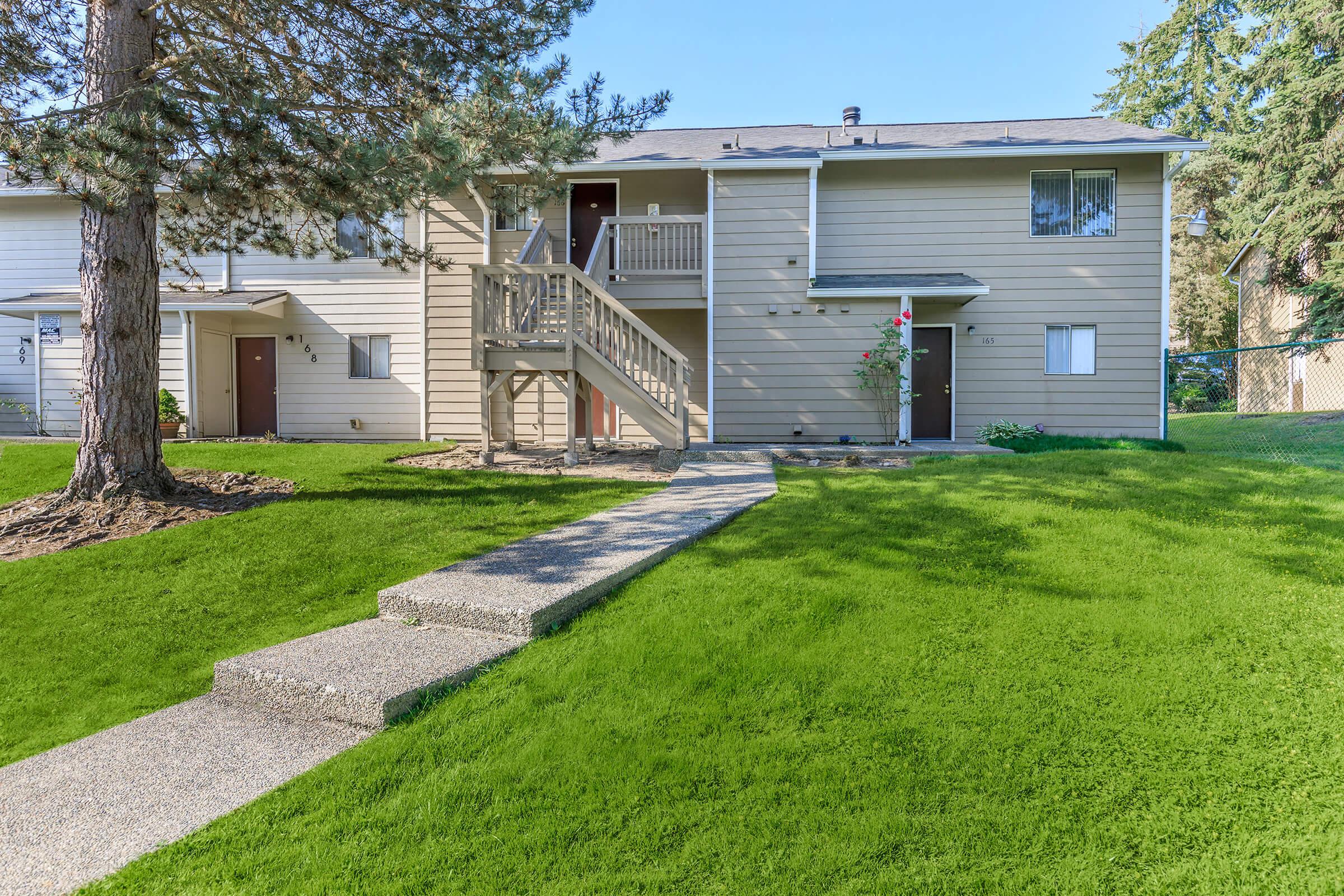 A two-story apartment building with a balcony on the second floor. A concrete pathway leads to the entrance, surrounded by lush green grass. Trees are in the vicinity, providing some shade. The units are painted in a light color, and stairs lead to the upper level.